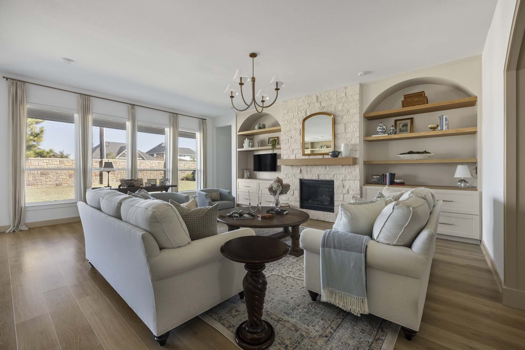 Living room with white sofas, a round coffee table, built-in white shelves, a fireplace, and large windows with curtains, overlooking a backyard.