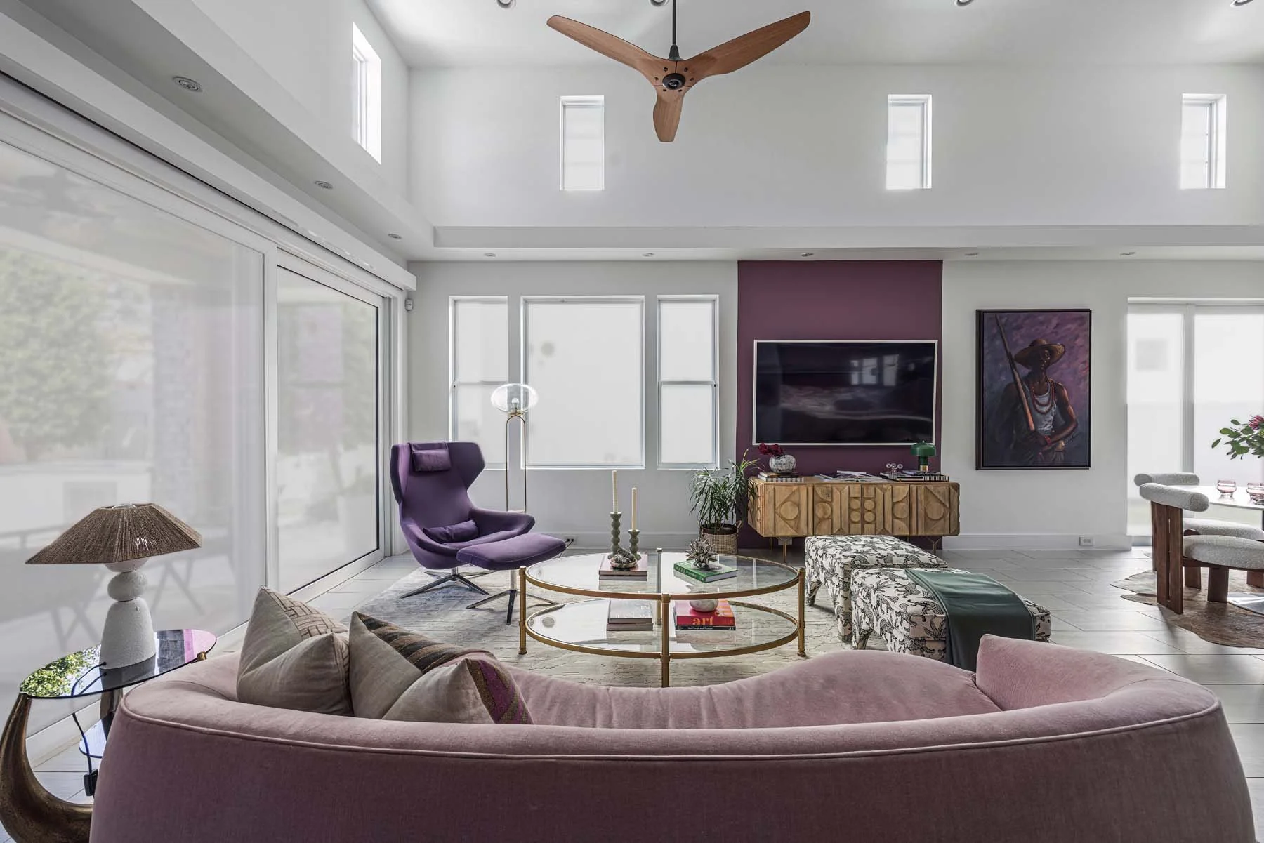 Modern living room with pink curved sofa, glass coffee table, purple lounge chair, patterned ottomans, wall-mounted TV, art portrait, potted plants, floor lamp, wooden sideboard, and ceiling fan.