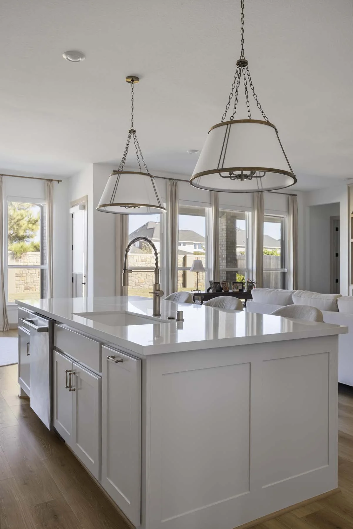 Modern kitchen with white island counter, double pendant lights, large windows, and a living room with a white couch in the background.