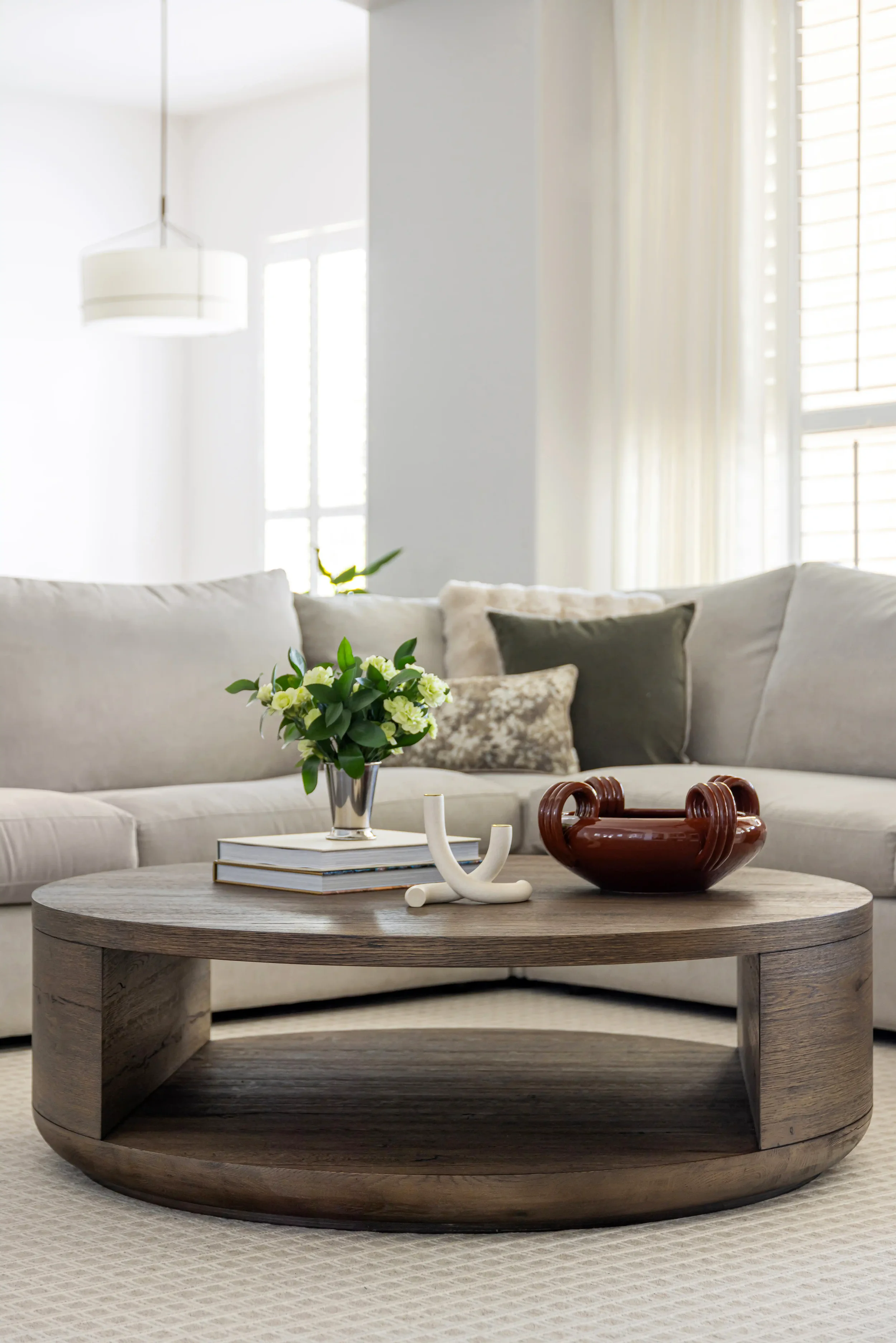 Living room with off-white sofa, decorated with pillows, a wooden coffee table with books, a flower vase, a decorative white object, and a ceramic bowl. Bright natural light coming through large windows with curtains.