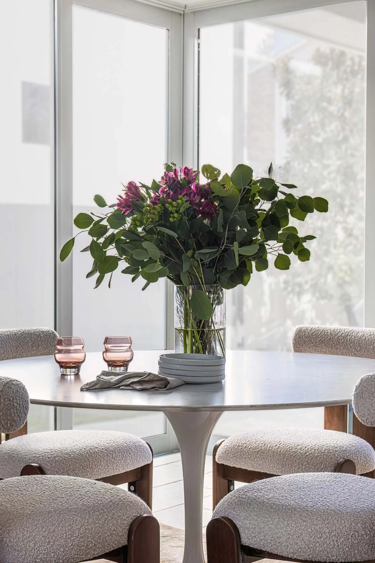 A round white table with a large bouquet of pink and purple flowers, green leaves, and eucalyptus in a clear glass vase. Four upholstered chairs with wooden legs surround the table. Two pink glasses and a cloth napkin are on the table. Large windows 