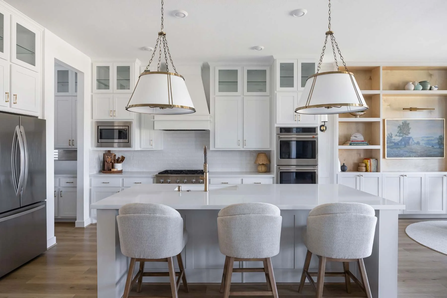 A modern kitchen with white cabinets, a large white island with bar stools, stainless steel appliances, and open shelving displaying dishes and decorative items.