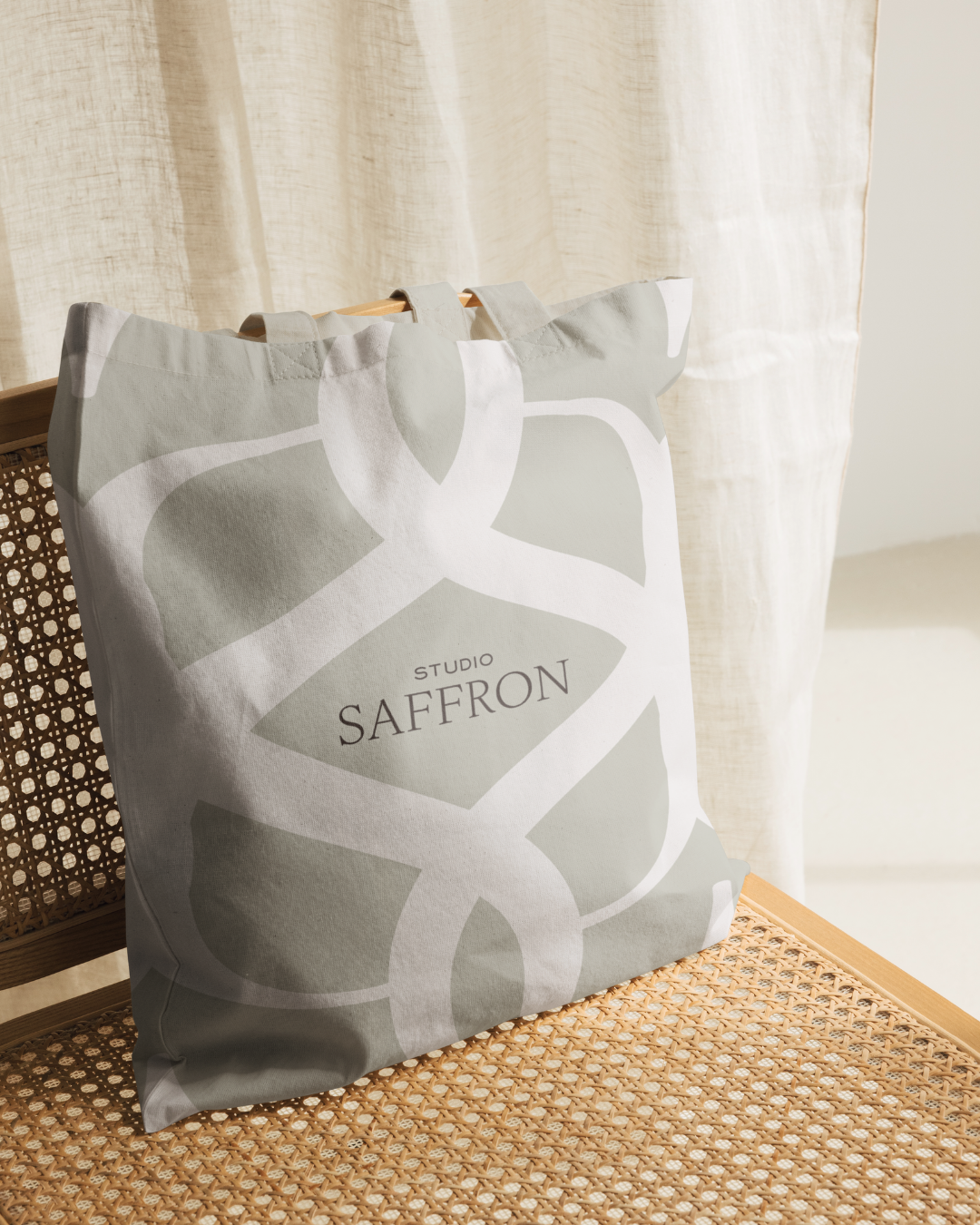 A fabric tote bag with a beige and white design and the words 'Studio Saffron' printed on it, resting on a wooden chair with a woven seat.
