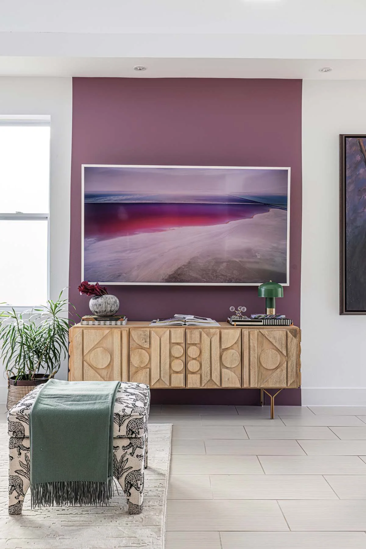 Living room with a purple accent wall, a large framed painting, a wooden sideboard, potted plants, a patterned stool with a green throw blanket, and a window with blinds.