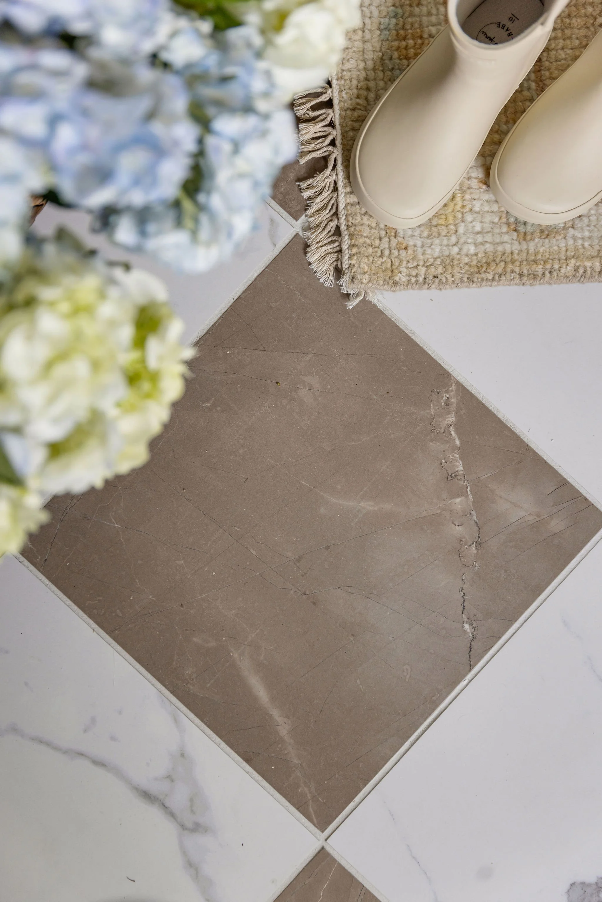 Corner of a floor with brown marble tiles and white marble tiles, beige textured rug, white rain boots on the rug, and a blurred bouquet of white and blue flowers in the foreground.
