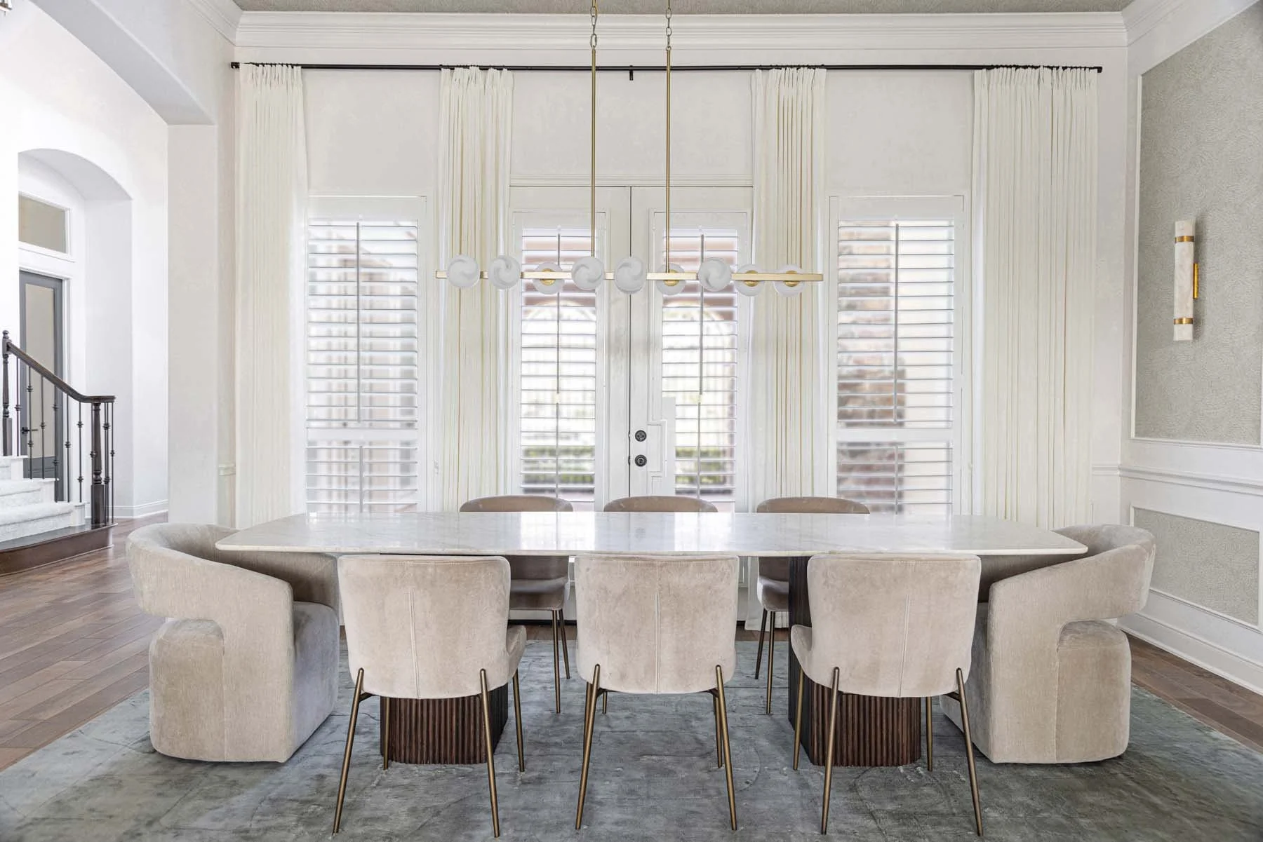 Modern dining room with a large oval white marble table, surrounded by ten beige upholstered chairs, set on a gray rug. Large windows with white shutters and cream-colored curtains allow natural light. A contemporary chandelier with globe lights hang