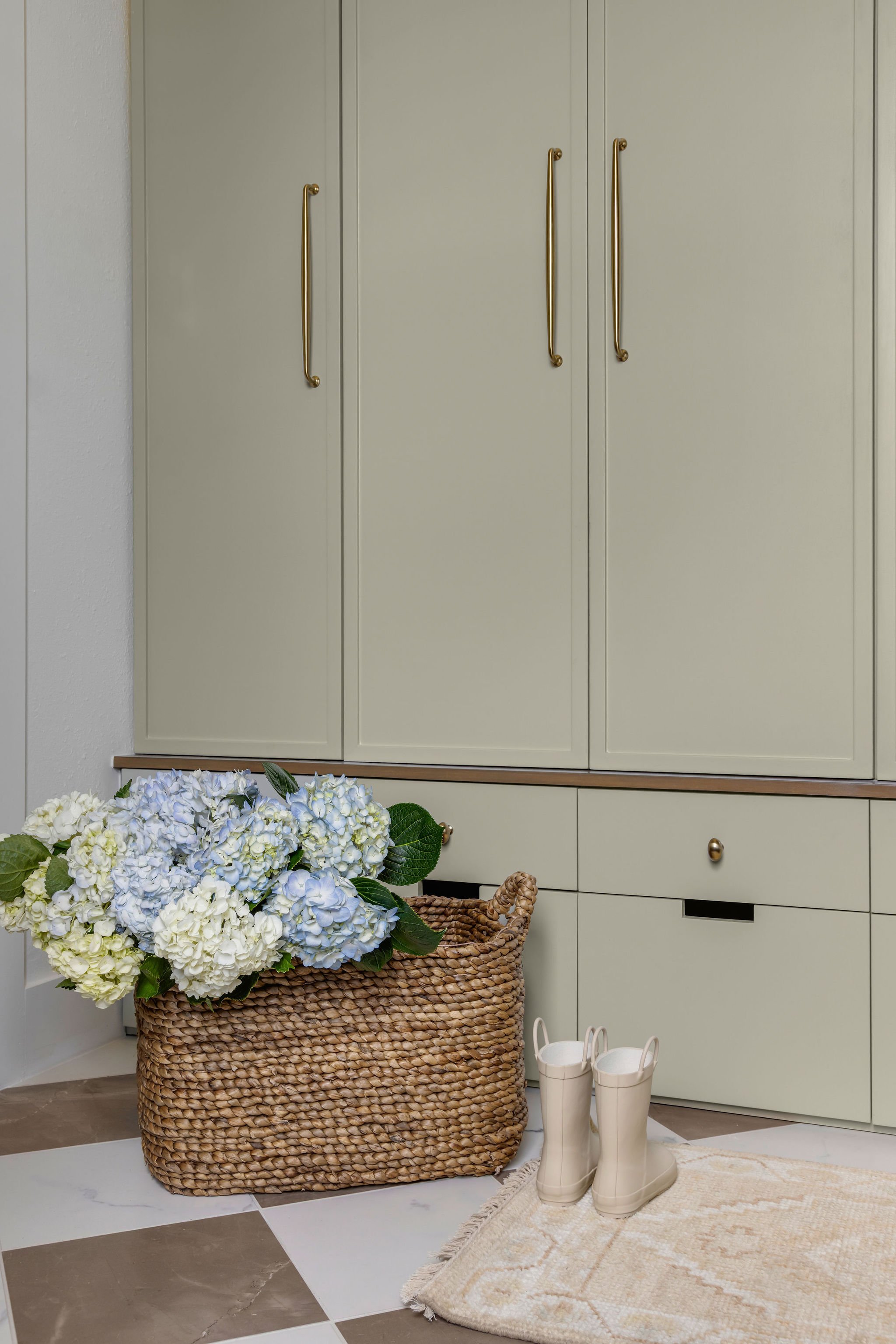A wicker basket filled with blue and white hydrangea flowers located on a tiled floor in front of pale green cabinets with brass handles. Next to the basket are a pair of white rain boots inside a beige rug.