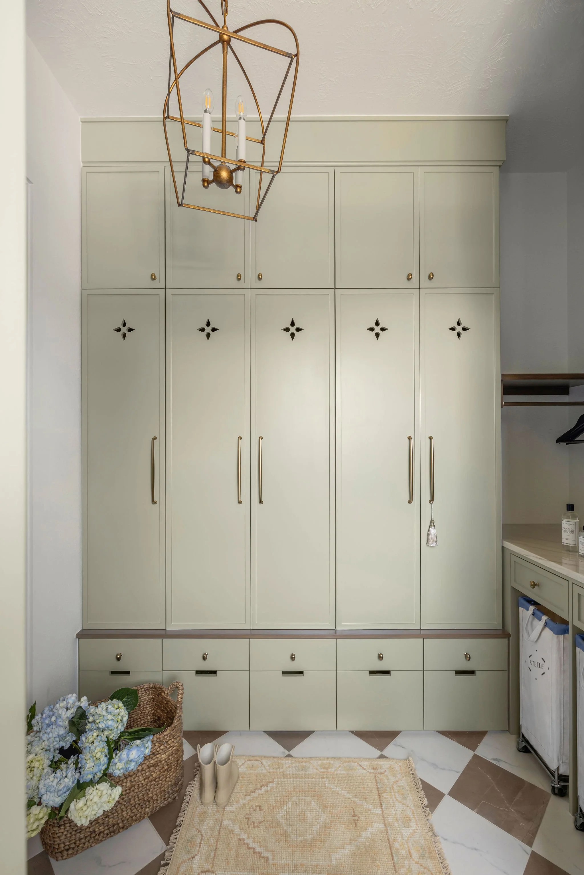 A bedroom corner corner with a built-in white wardrobe with gold handles. To the right, a small white shelf with a glass door and clothes hanging inside. On the floor, a beige and cream rug, white shoes, a woven basket with blue hydrangeas, and a sma