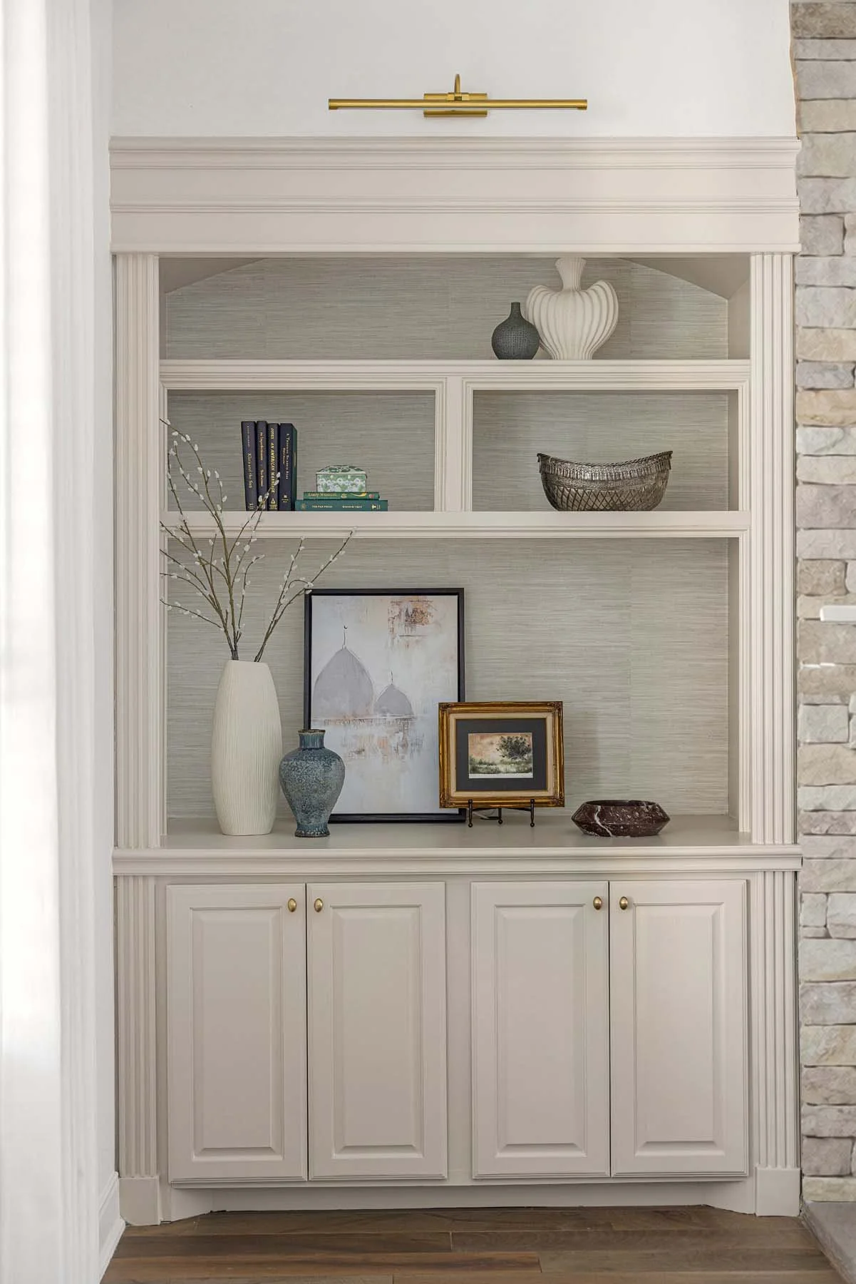 Built-in white bookshelf with decorative items, including vases, framed pictures, books, and a basket, against a light-colored wall and stone wall on the side.
