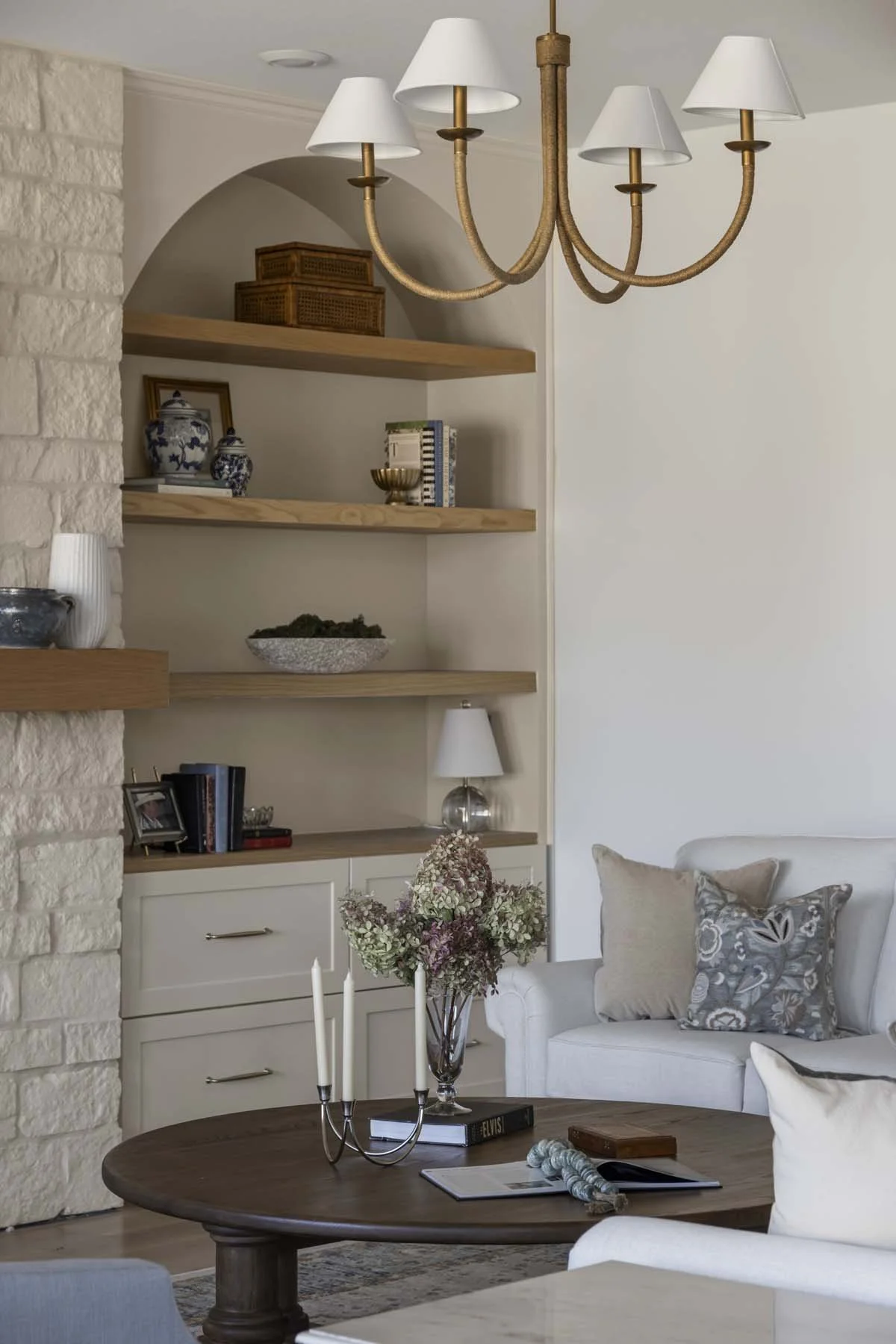 Living room with a chandelier, open wooden shelves with decorative items, a white armchair with pillows, a wooden coffee table with books, a candle holder, and a vase with flowers.