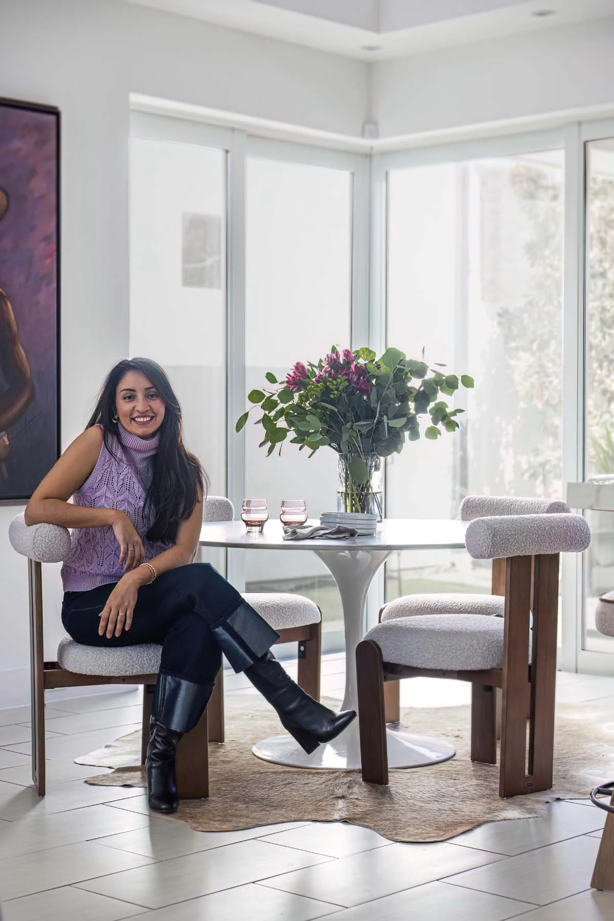 A woman sitting at a round dining table with a large bouquet of pink and purple flowers, in a bright room with large windows.