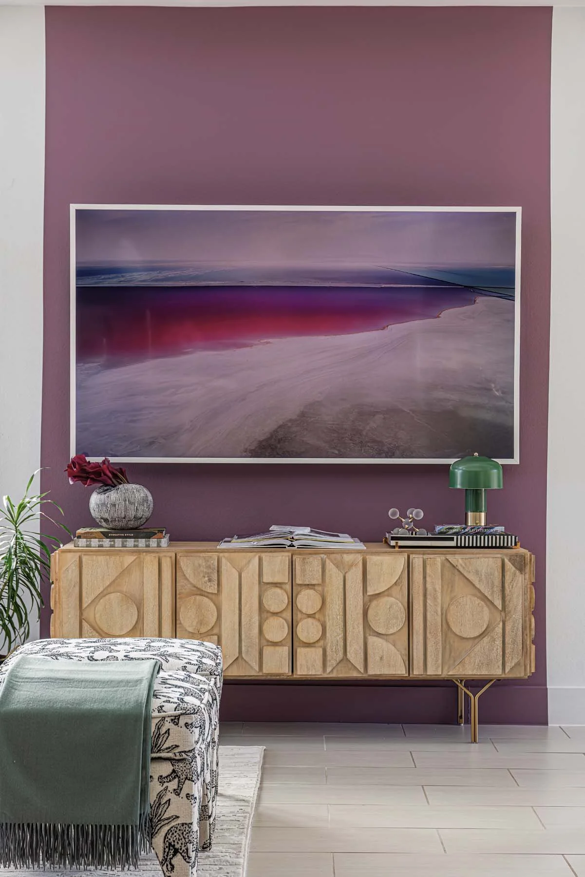 Interior of a stylish living room with a purple accent wall, a large abstract landscape painting, a wooden sideboard, a green table lamp, a decorative vase with red flowers, and a patterned ottoman.