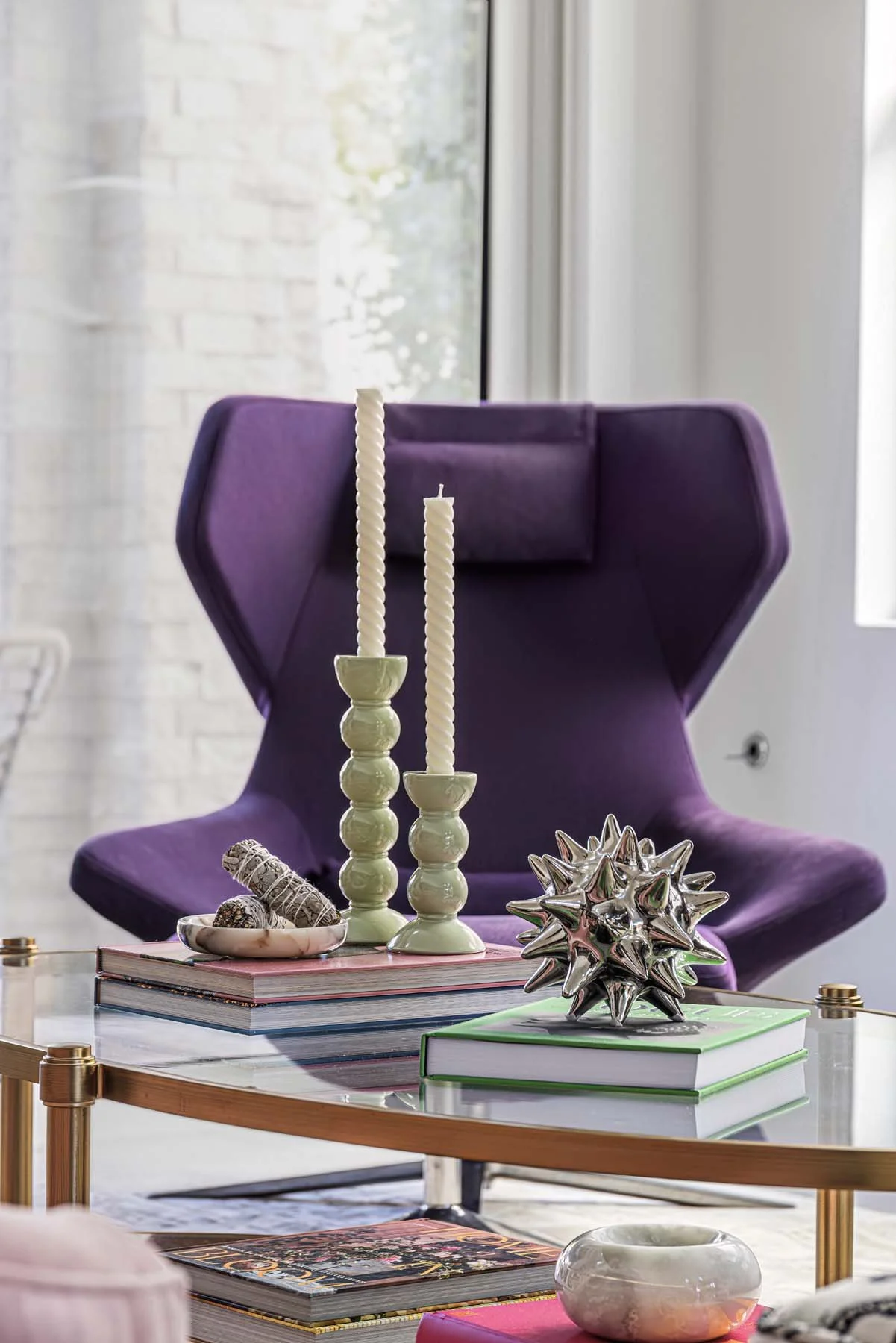 Decorative table with candles, books, and a metallic sculpture in front of a purple armchair near a window.