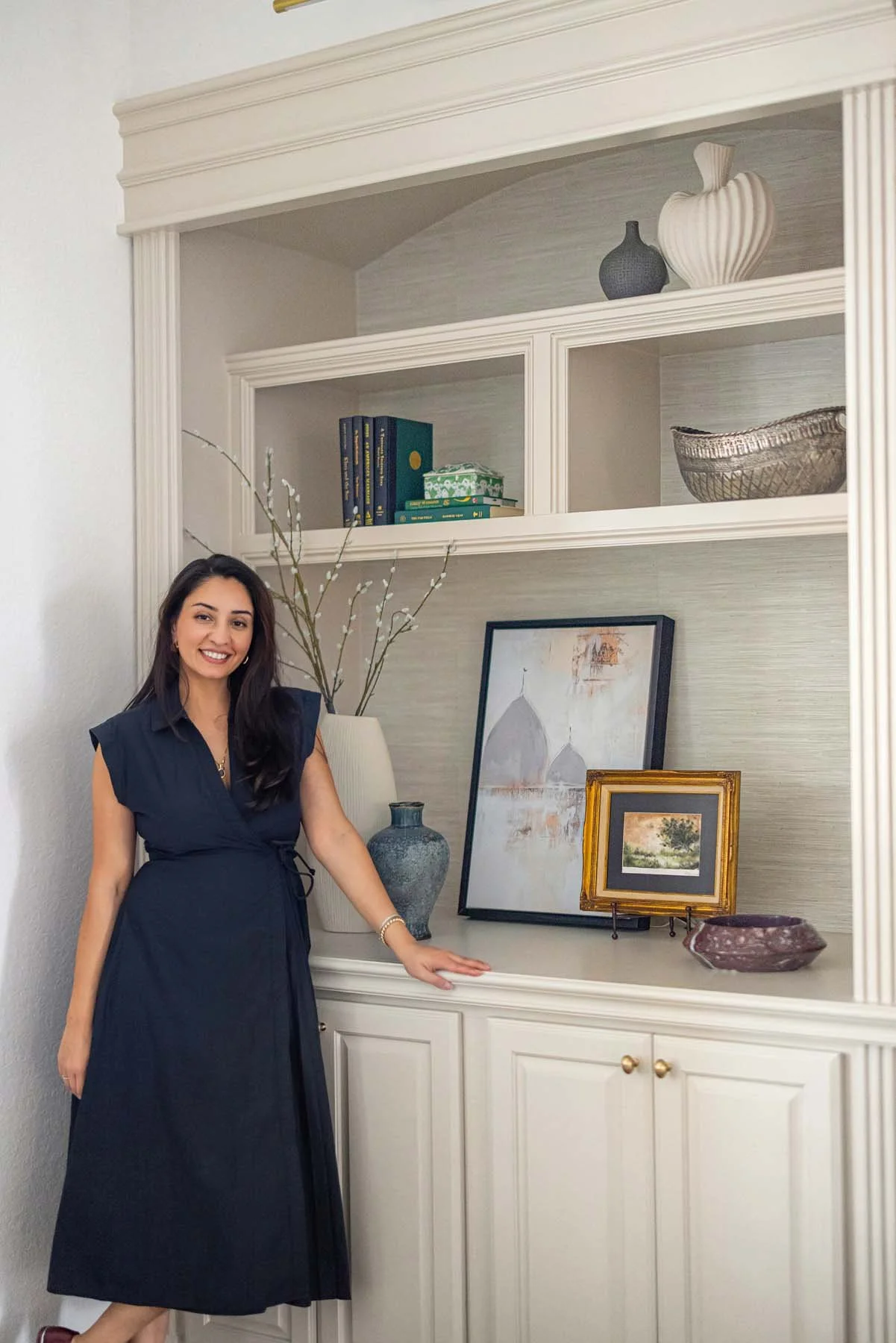 A woman in a navy blue dress standing next to a white built-in bookshelf, smiling at the camera.