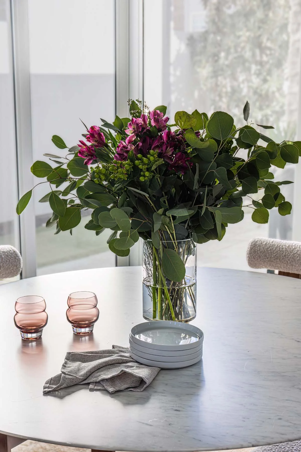 A white marble table with a large glass vase filled with purple and green flowers and foliage, two pink glasses, white bowls, and a gray cloth, in front of large glass sliding doors showing trees outside.