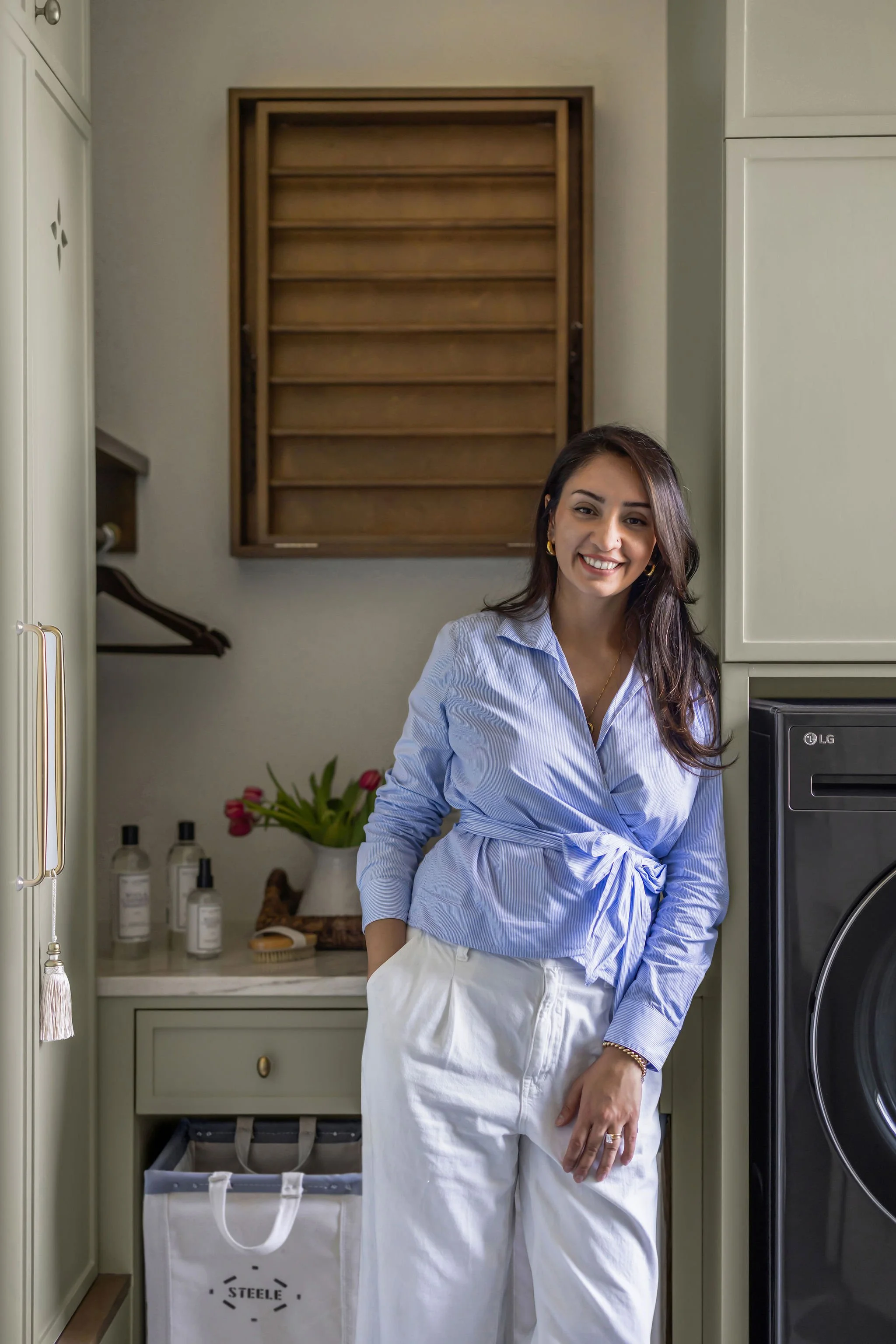 A woman smiling in a laundry room, wearing a light blue shirt and white apron, standing next to a washing machine with laundry baskets and shelves with bottles and flowers.