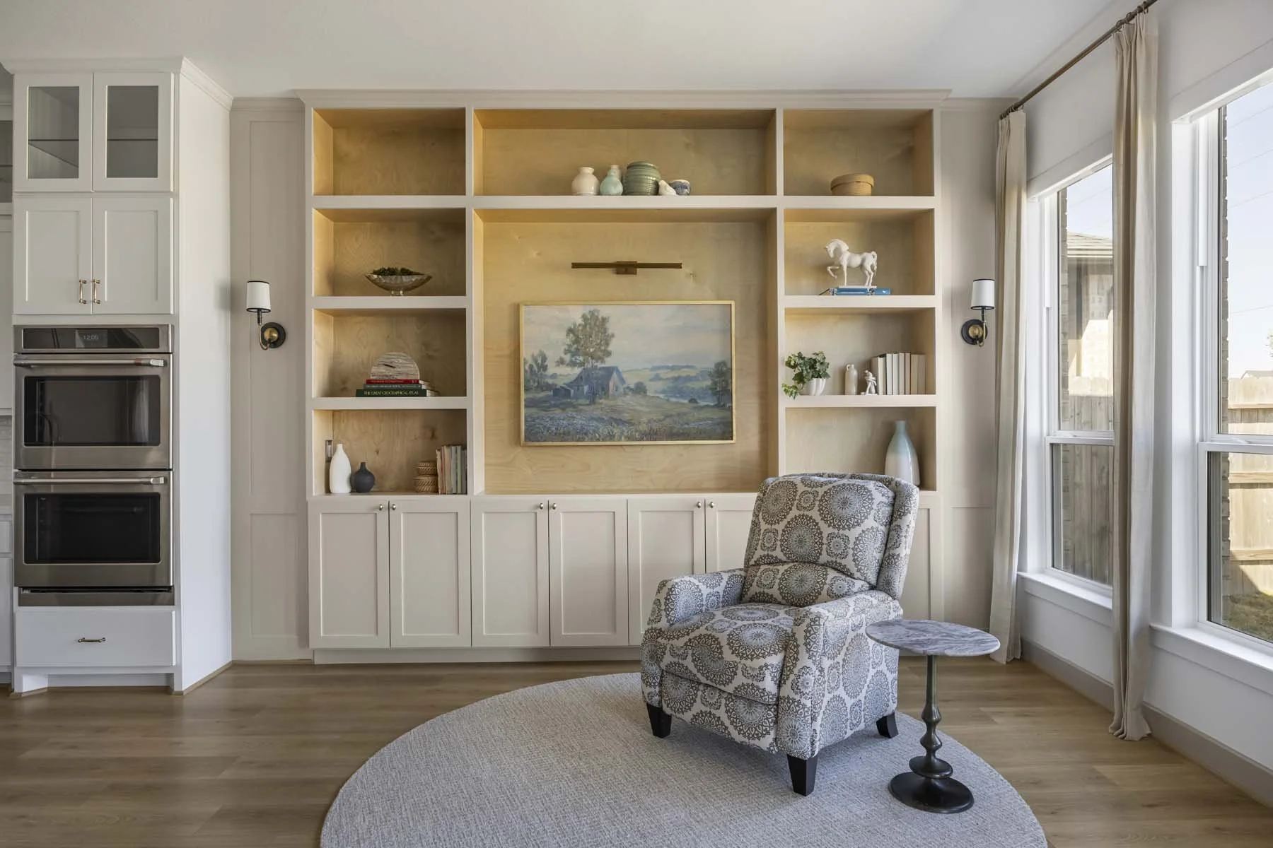 Living room with a white built-in bookshelf, a framed painting of a countryside landscape, a patterned armchair, a small side table, and large windows with curtains.