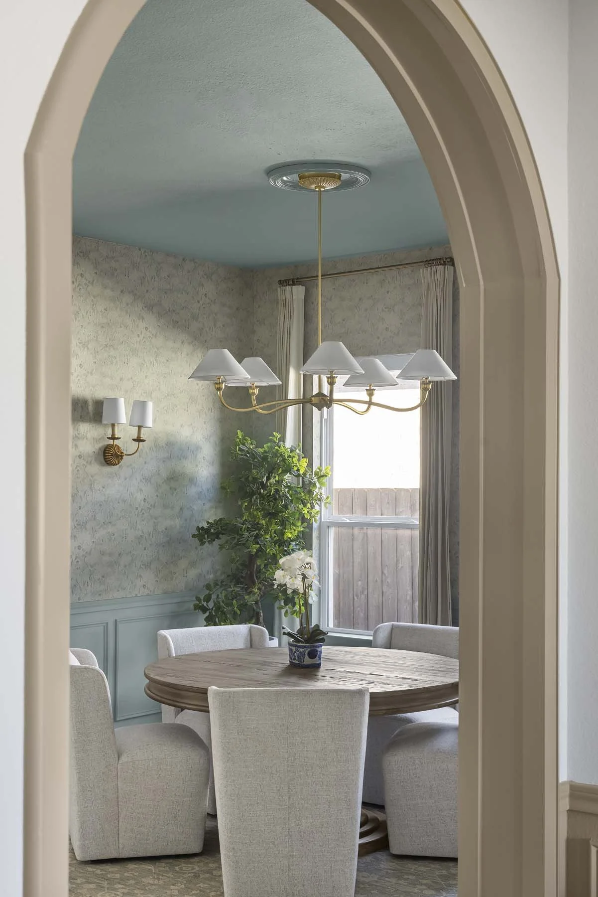 Dining room with a round wooden table, six upholstered chairs, a chandelier, a window with curtains, a potted orchid, and a tall plant, viewed through an arched doorway.