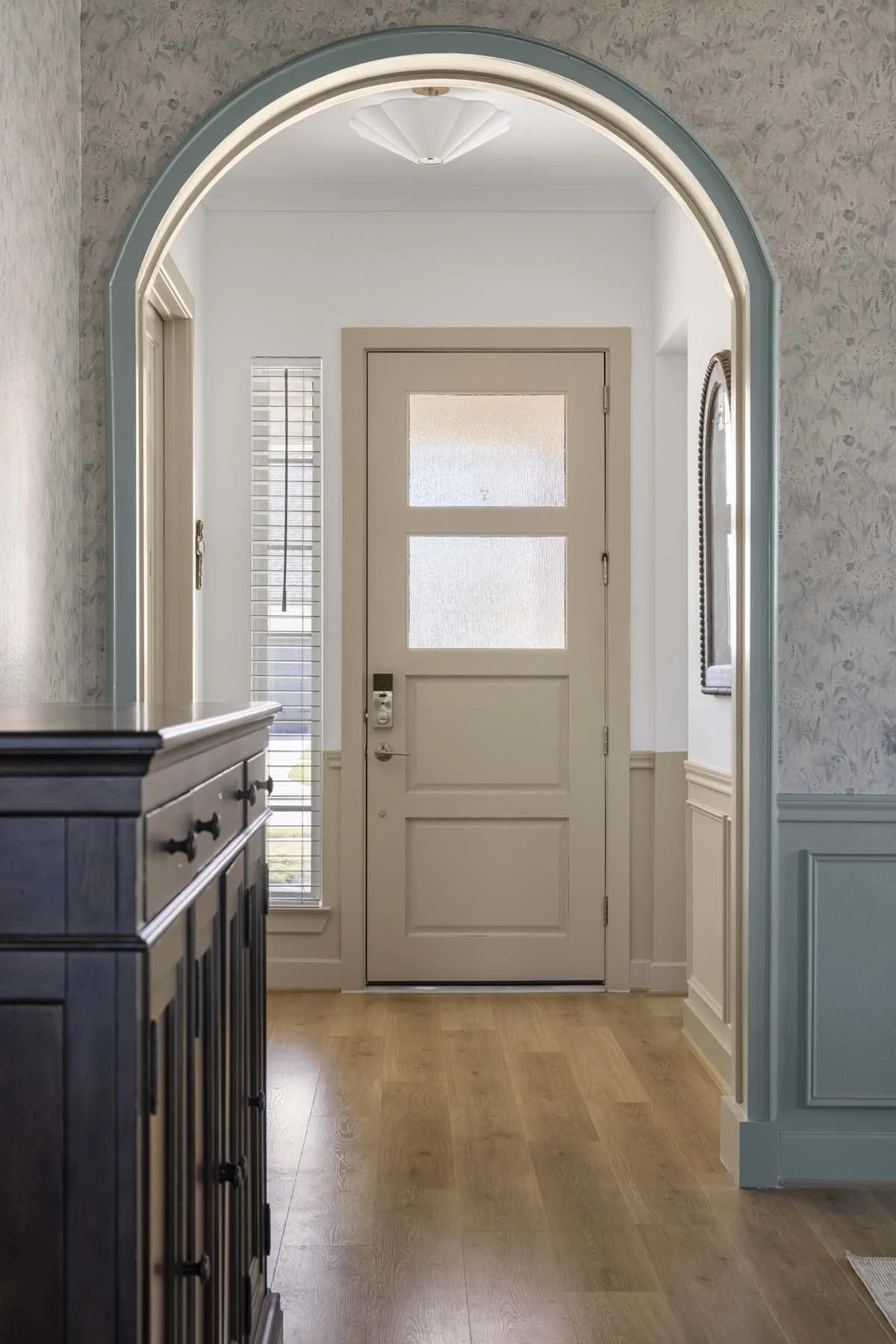 Entryway with a closed door with frosted glass panels, surrounded by white walls with crown molding, a black console table on the left, and a mirror on the right. There are tall windows with blinds on either side of the door, and hardwood flooring.