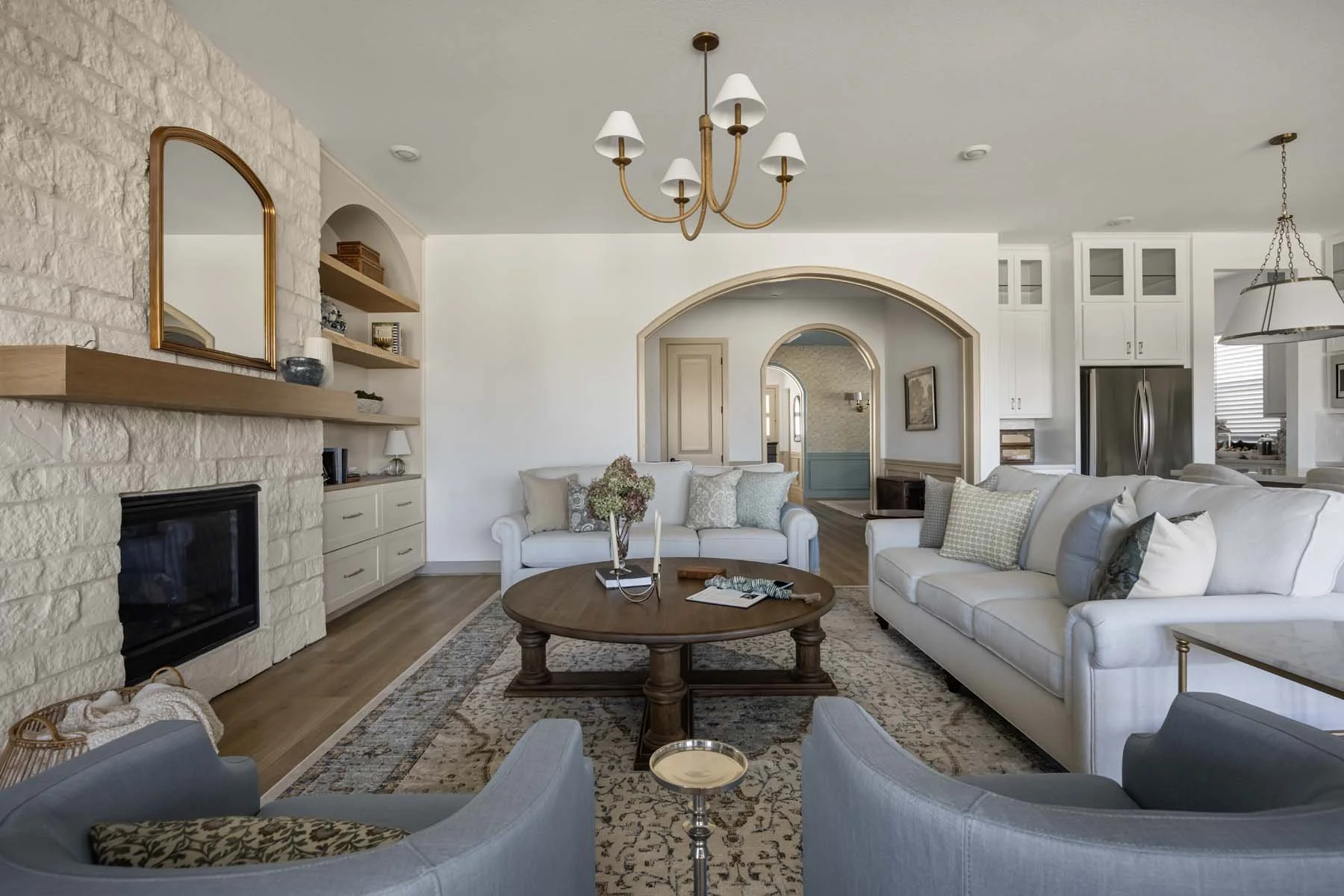 Bright living room with white and gray sofas around a wooden coffee table, a white brick fireplace with a wooden mantel, a chandelier with white lampshades, built-in shelves, a patterned area rug, and open kitchen with white cabinets and stainless st