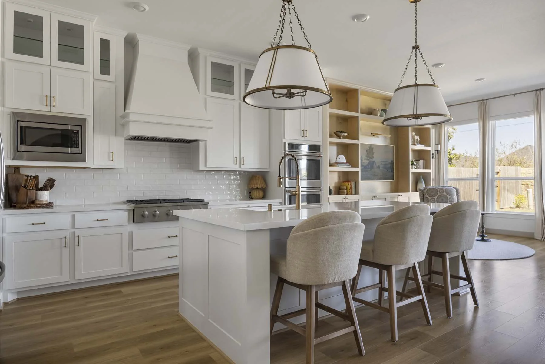Bright open kitchen with white cabinets, a central island with beige upholstered chairs, and a living space with large windows and beige curtains in the background.