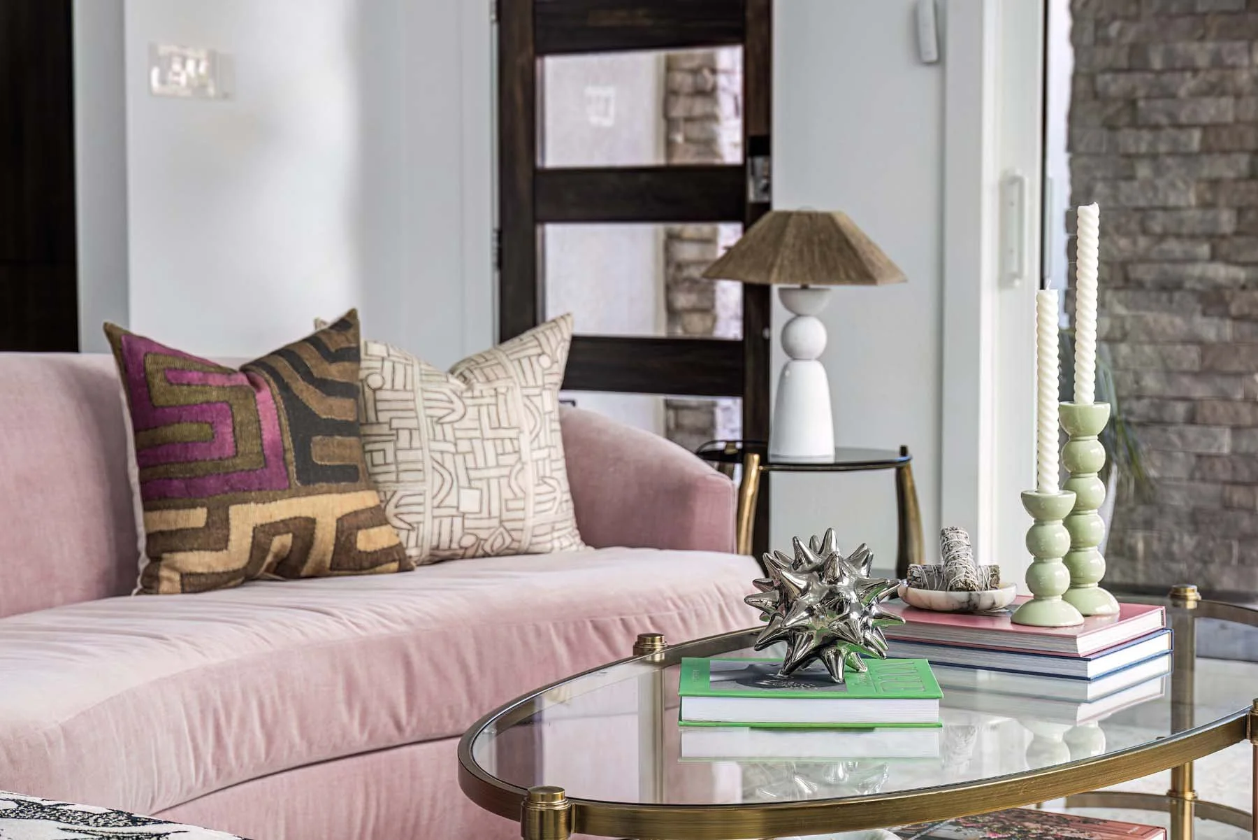 Living room with pink velvet sofa, patterned throw pillows, gold and glass coffee table, decorative objects including spiky silver sculpture and candlesticks, lamp with a beige shade, and a window with stone wall visible outside.