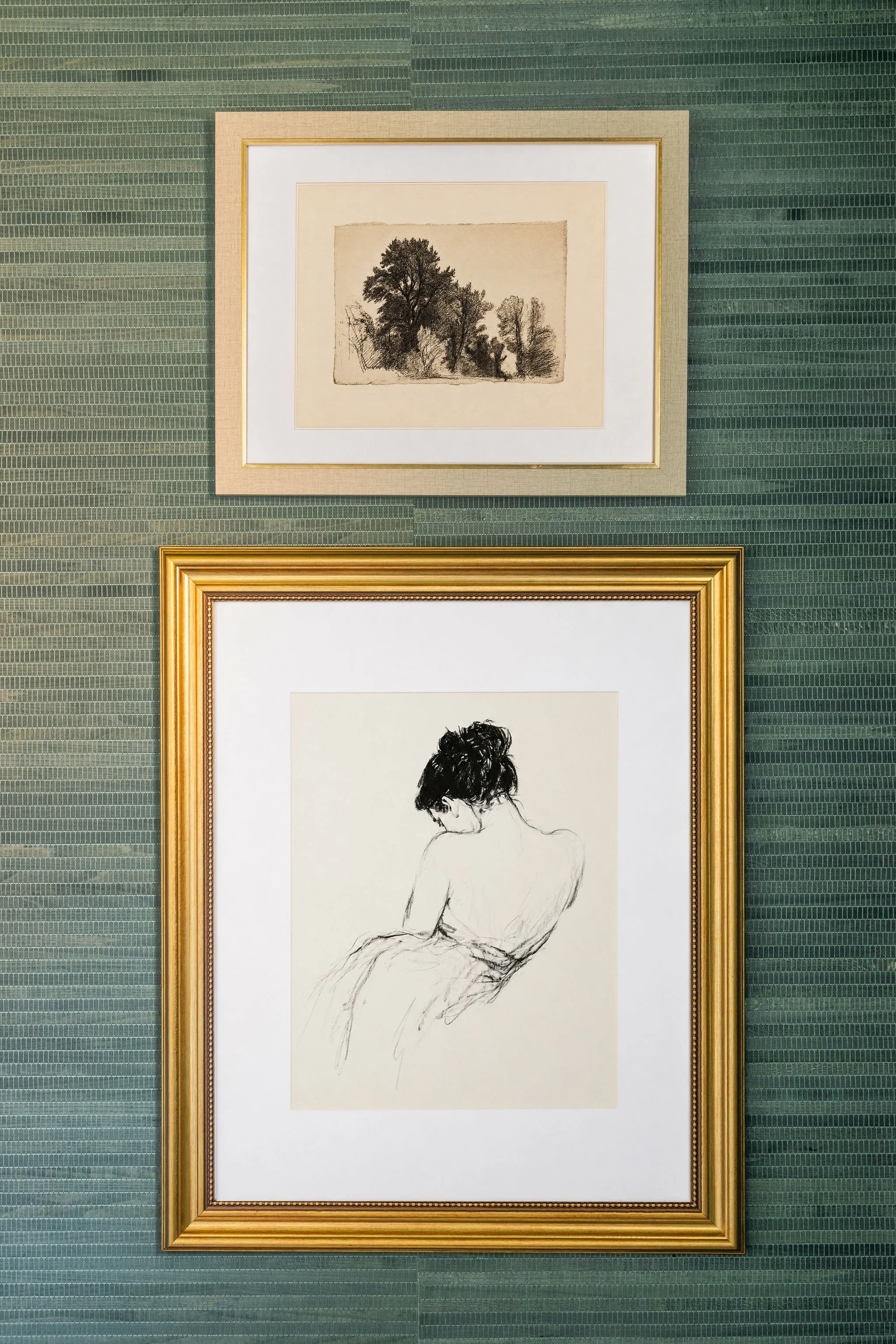 Two framed artworks on a textured green wall, the top one depicts a sketch of trees, the bottom one is a drawing of a woman with short hair, sitting and viewed from the back.