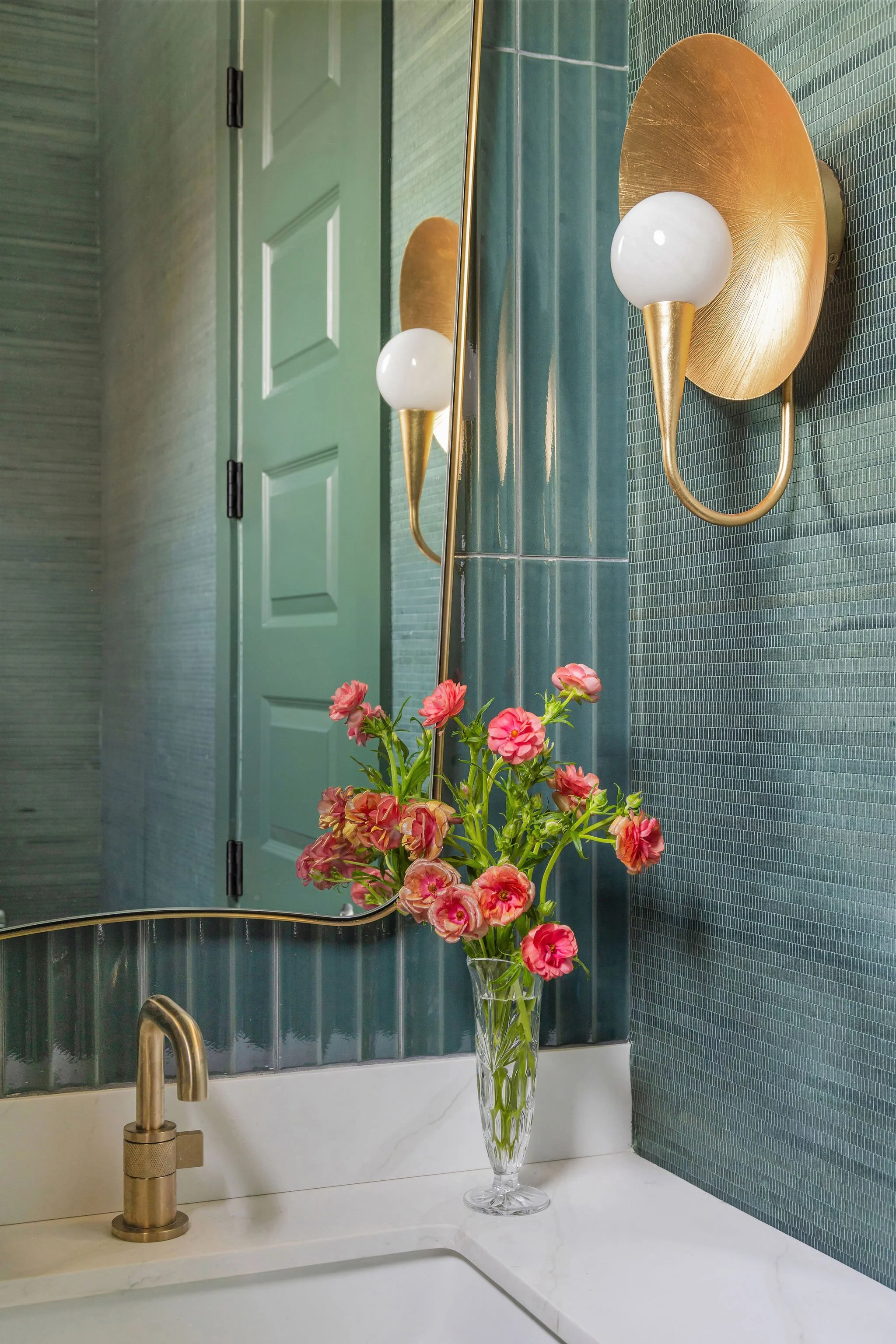 A bathroom sink area with a gold faucet, a large mirror, green textured tiled wall, a green door, a glass vase with pink flowers, and a gold wall sconce.