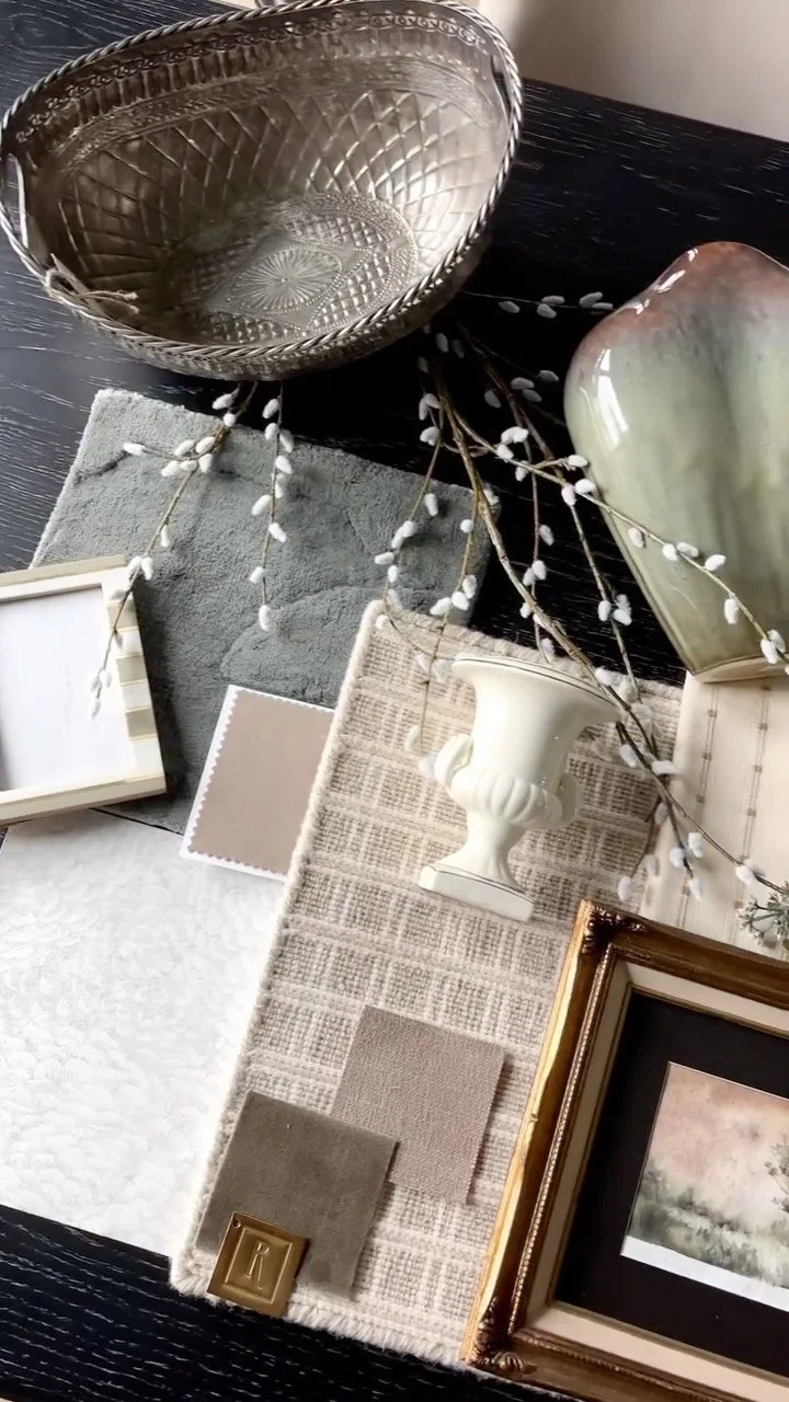 Decorative objects including a silver bowl, a ceramic vase, a white pedestal vase, a small picture frame, a beige and white color palette fabric, fabric swatches, and a framed artwork on a dark wood surface.