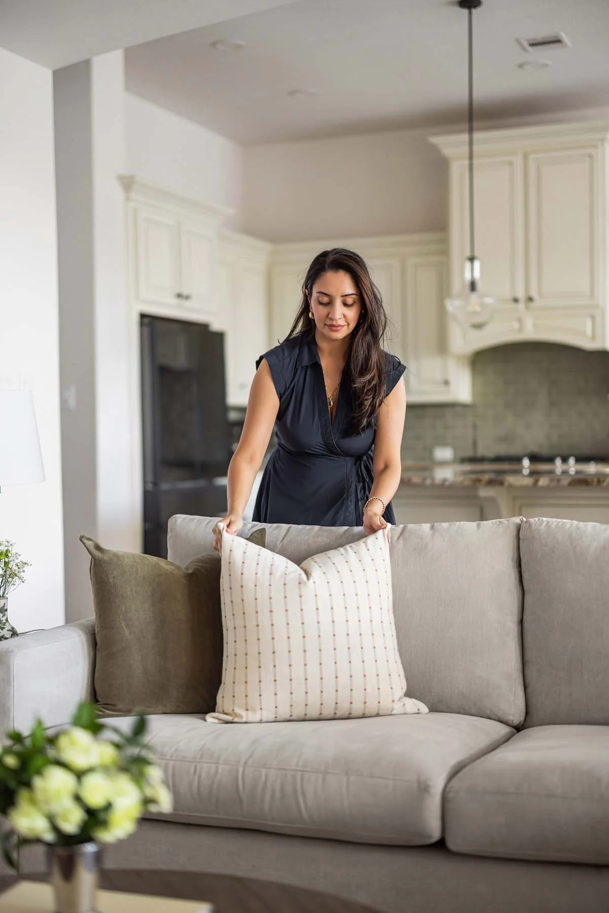 A woman in a black dress arranging cushions on a beige sofa in a living room with cream-colored cabinets and a kitchen in the background.