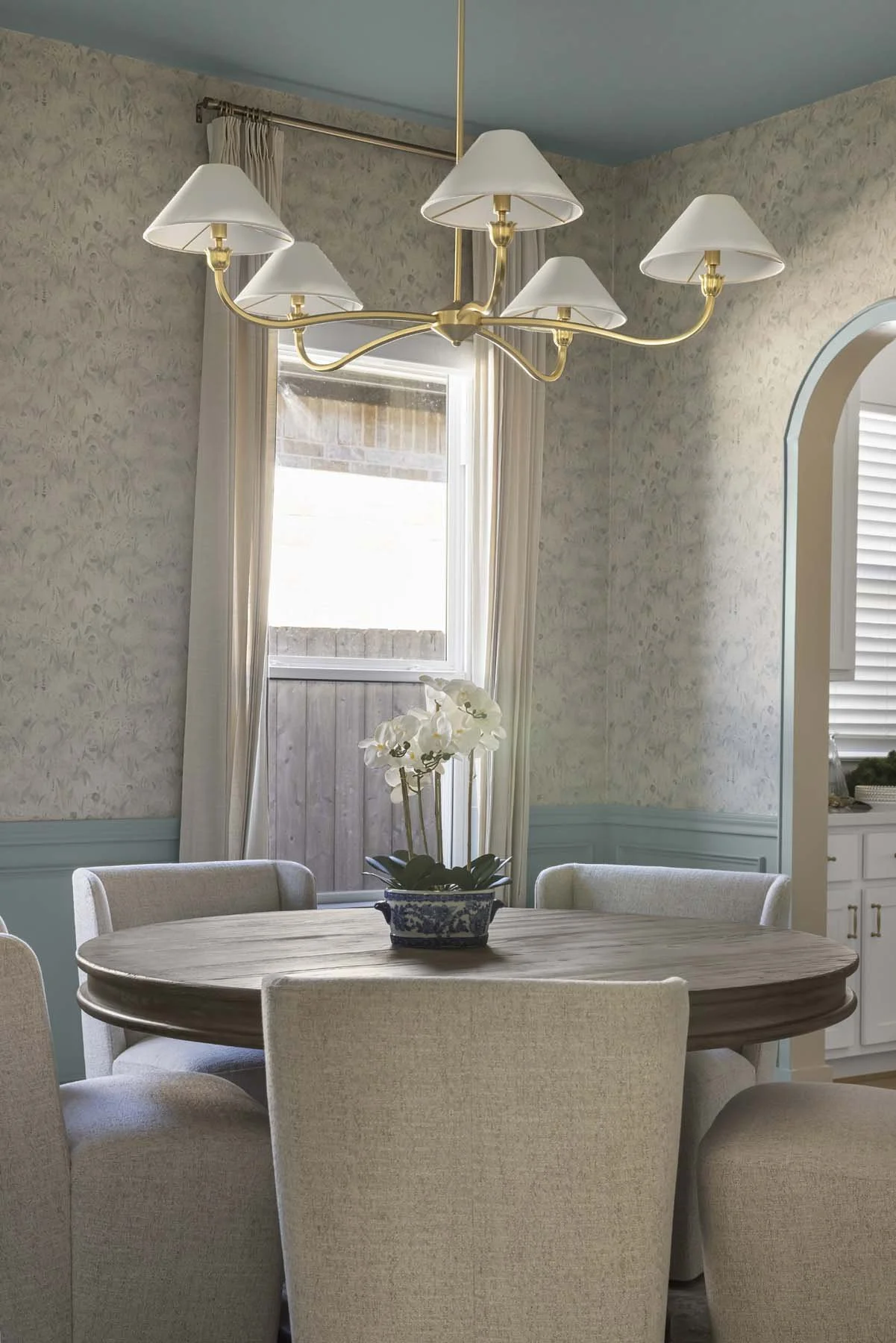 A dining room with a wooden round table, beige upholstered chairs, a white orchid plant in a blue and white ceramic pot, a window with white curtains, and a gold chandelier with five lampshades.