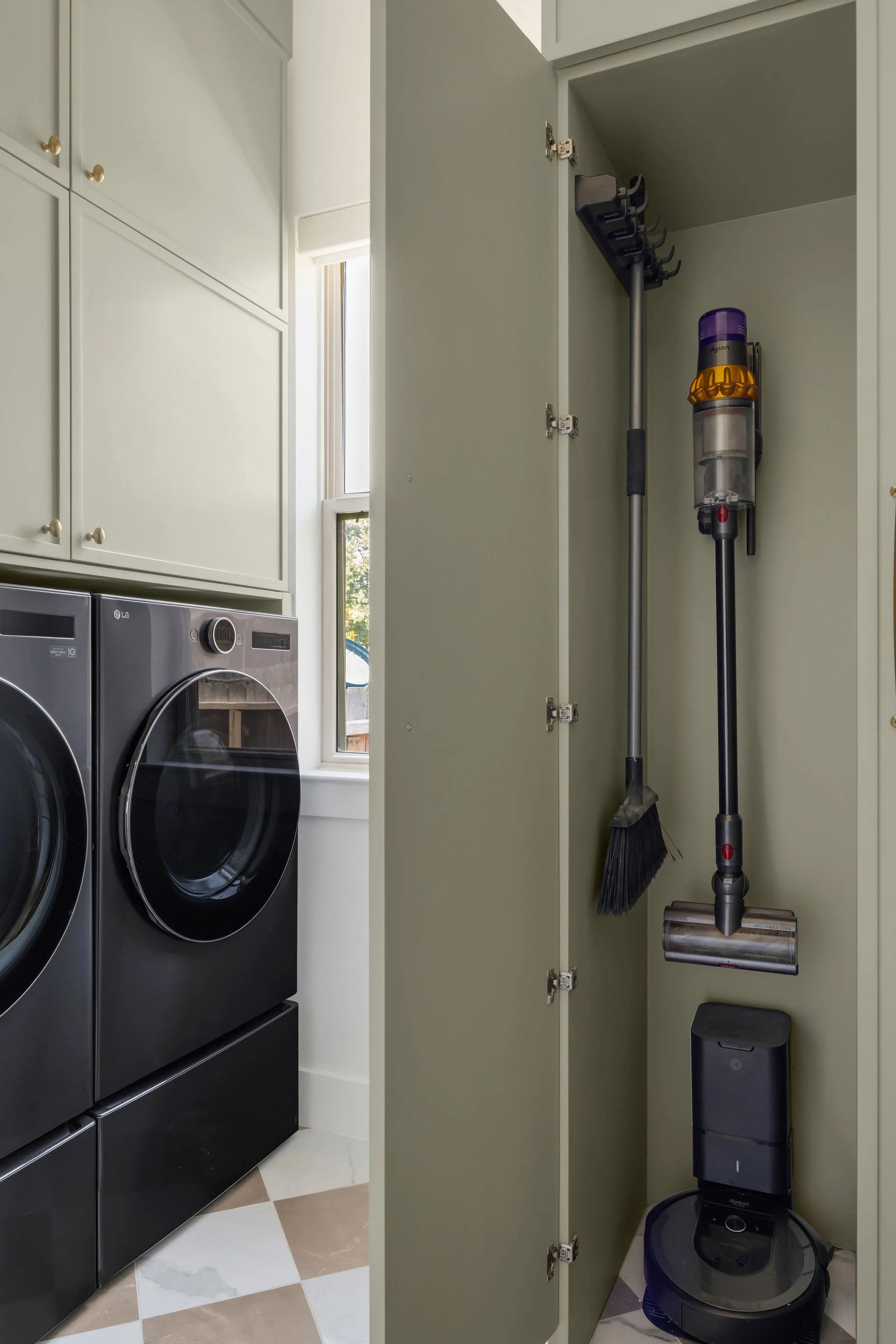 A laundry room with a front-loading washer and dryer next to each other, with open cabinet doors revealing cleaning tools like a broom, vacuum cleaner, and robot vacuum on the floor.