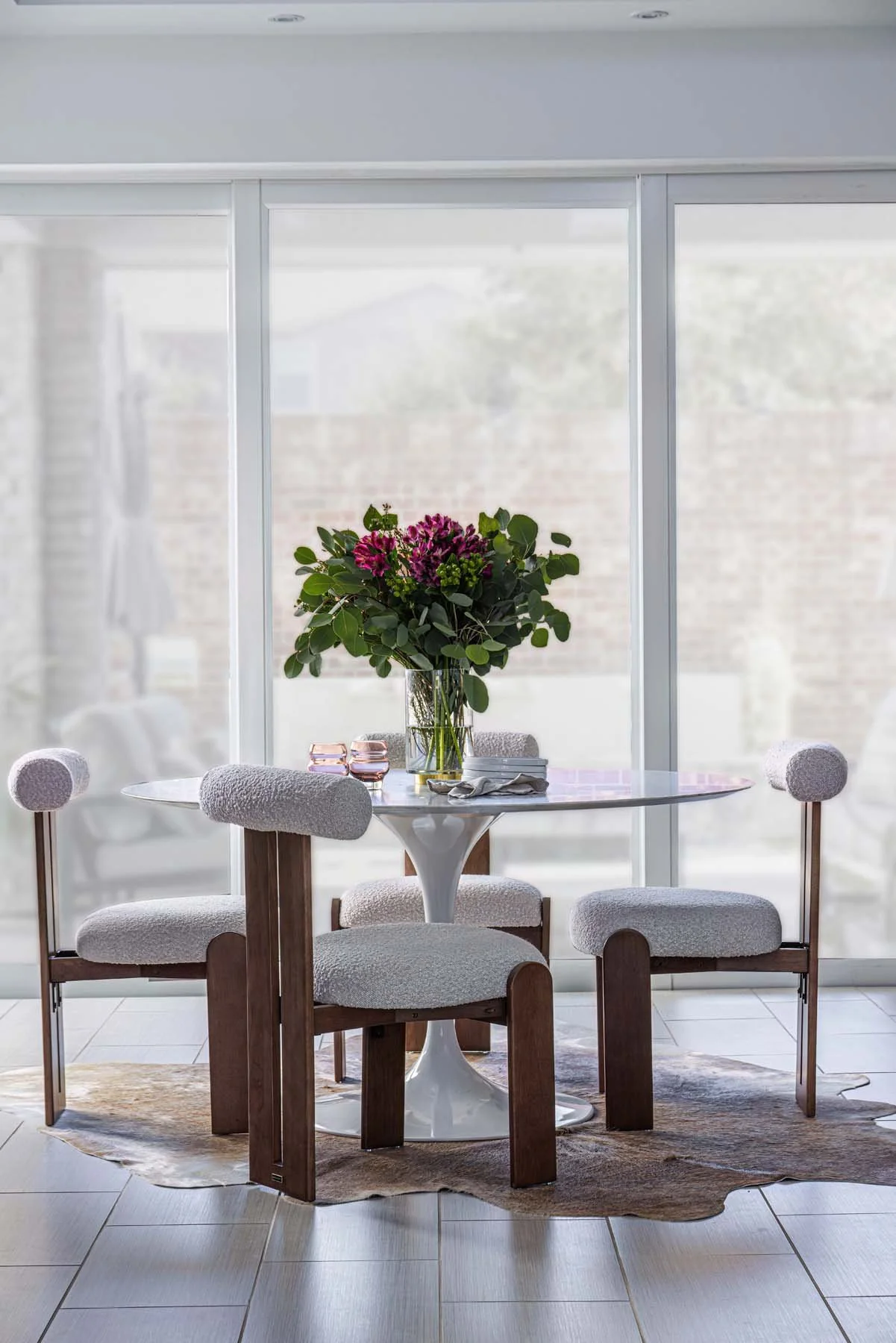 A round dining table with a white tulip base, topped with a large bouquet of purple and pink flowers, surrounded by four chairs with wooden legs and white upholstered cushions, placed on a beige rug near large sliding glass doors in a bright room.