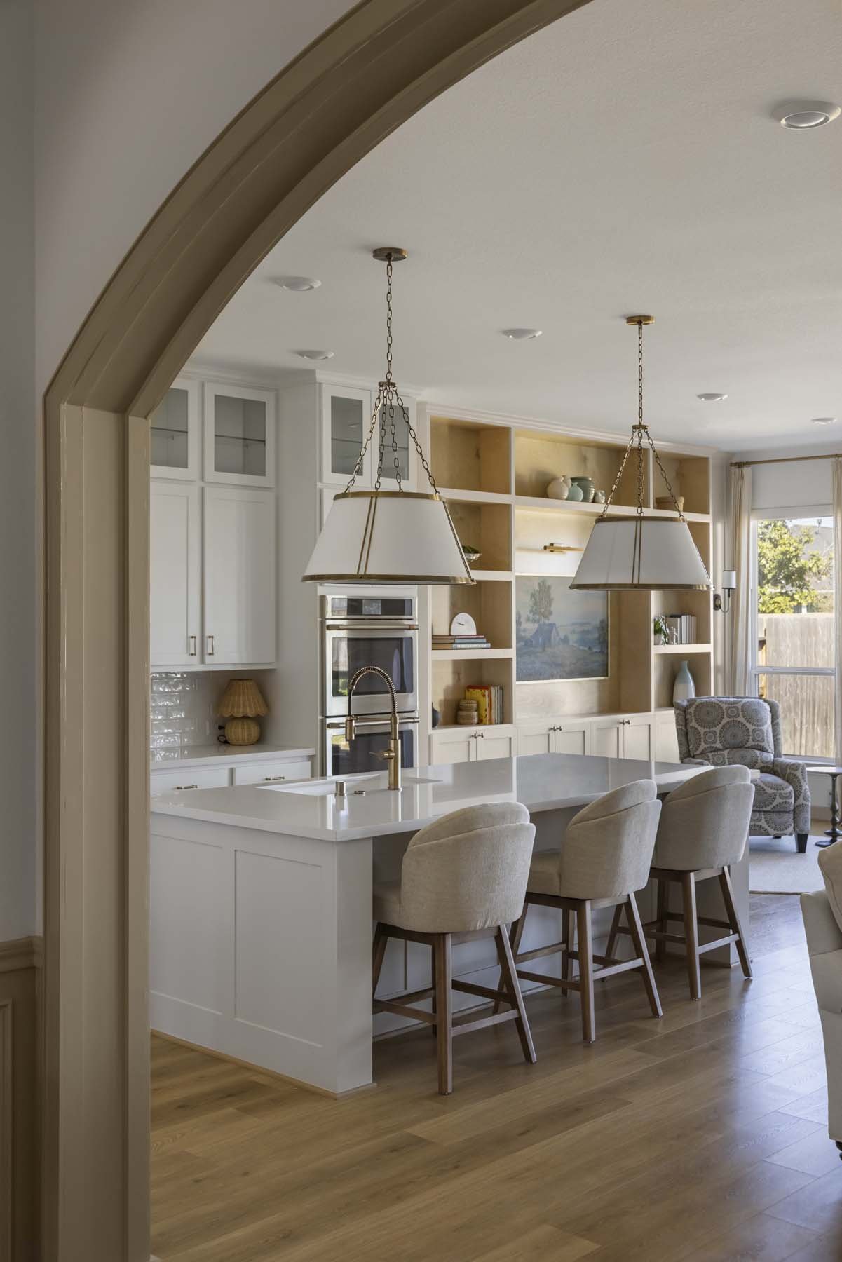 A modern kitchen and living area seen through an arched doorway. The kitchen features a white island with a gold faucet, beige barstools, and white cabinets. In the background, there are open shelves, a wall-mounted oven, and a window with a view of 