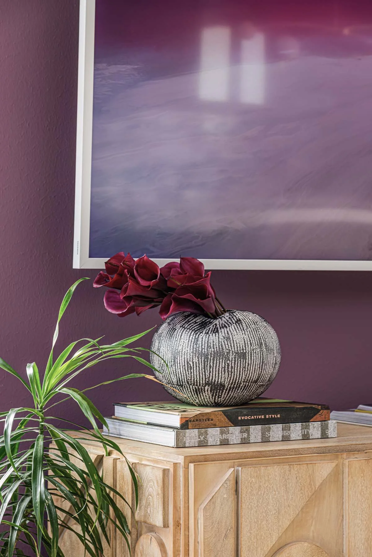 A decorative plant with green leaves in the foreground, a round patterned vase with dark red calla lilies on top, resting on a stack of books on a light wooden cabinet, with a purple wall and a large framed abstract artwork in the background.