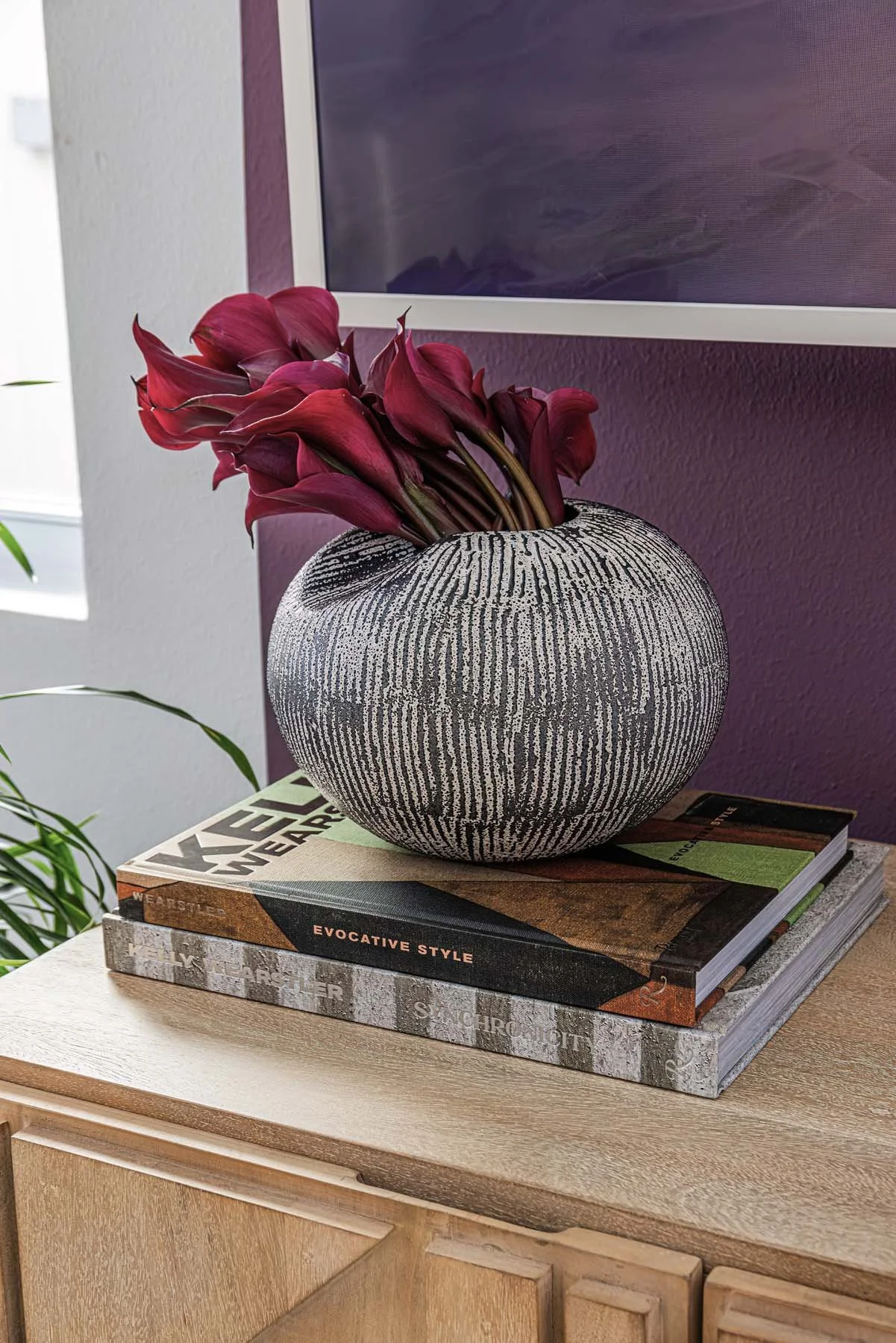 A decorative vase with a black and white textured pattern, holding dark red calla lilies, placed on top of two stacked art or design books on a wooden table.