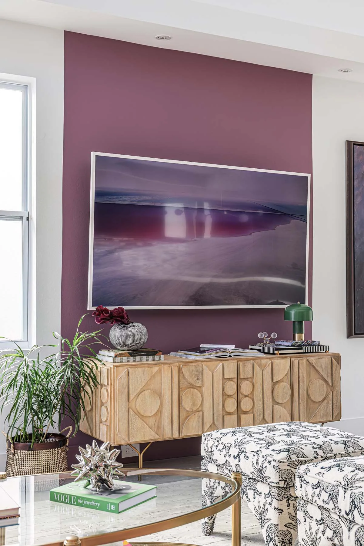 Living room with a purple accent wall, a flat-screen TV mounted on the wall, a wooden sideboard with geometric carvings, a green table lamp, a vase with flowers, books, and an abstract patterned upholstered chair and a glass coffee table with decorat