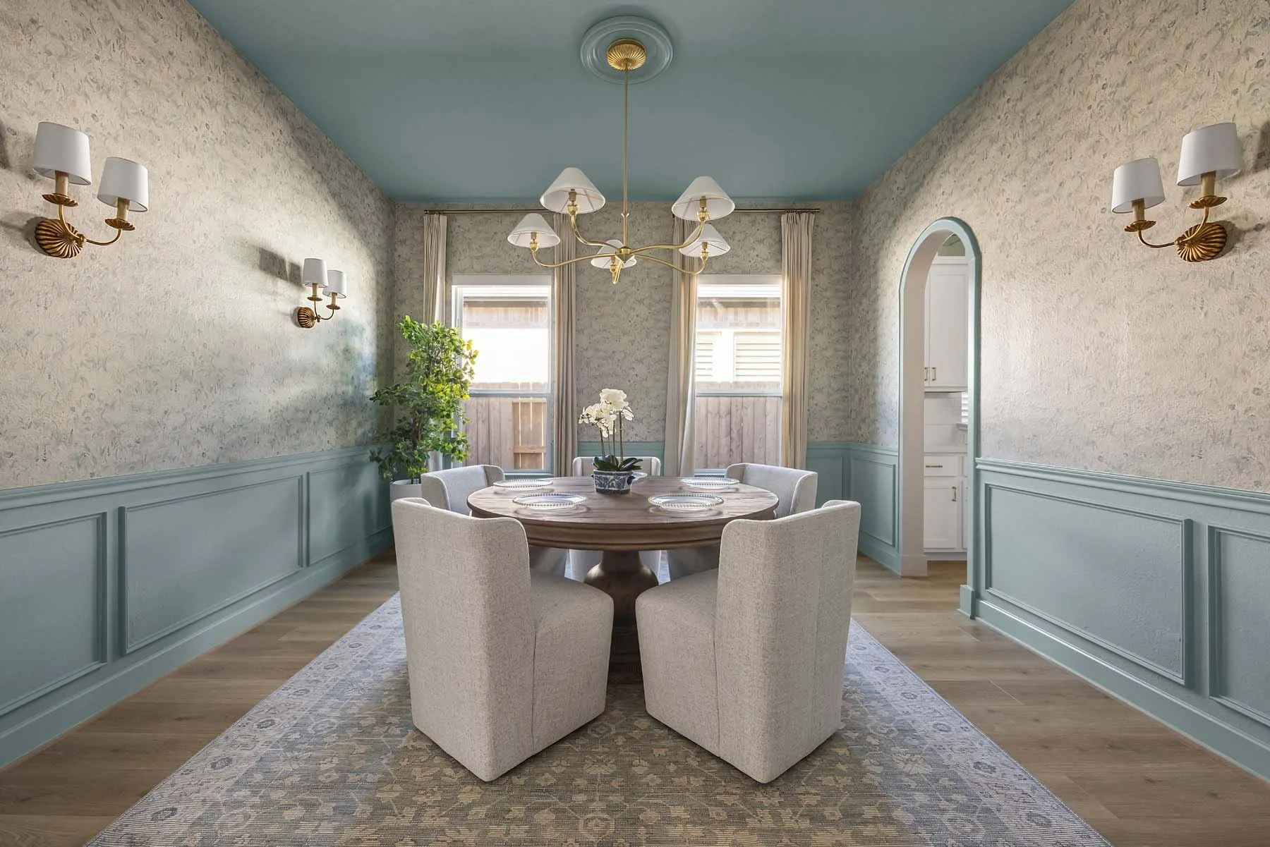 Dining room with round wooden table, eight beige upholstered chairs, floral centerpiece, patterned rug, light blue wainscoting, textured beige walls, hanging chandelier, wall sconces, windows with curtains, and an adjacent pantry area.