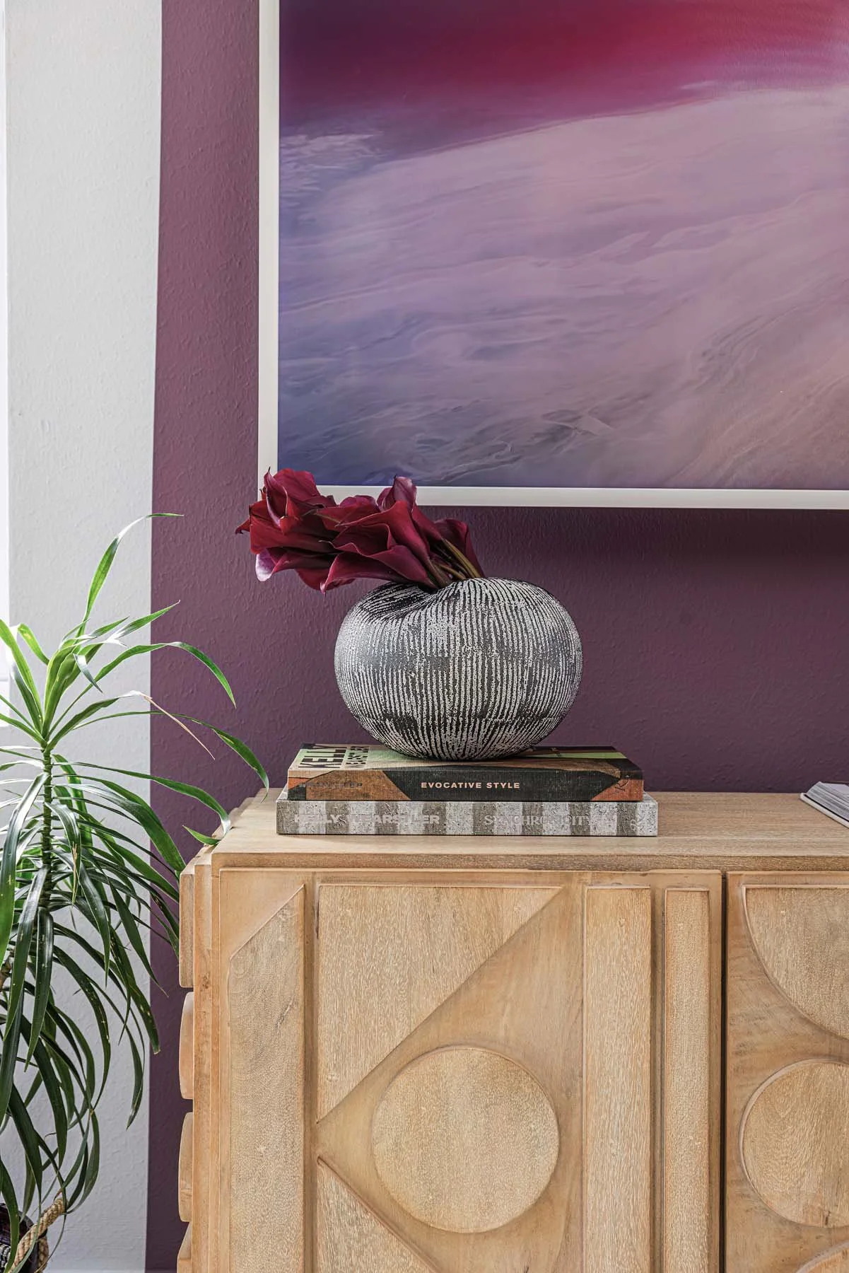 A decorative black and white ceramic vase with a textured pattern, holding dark red flowers, is placed on a stack of books on a light wooden sideboard. A green plant is to the left and a colorful abstract painting is on the purple wall behind.