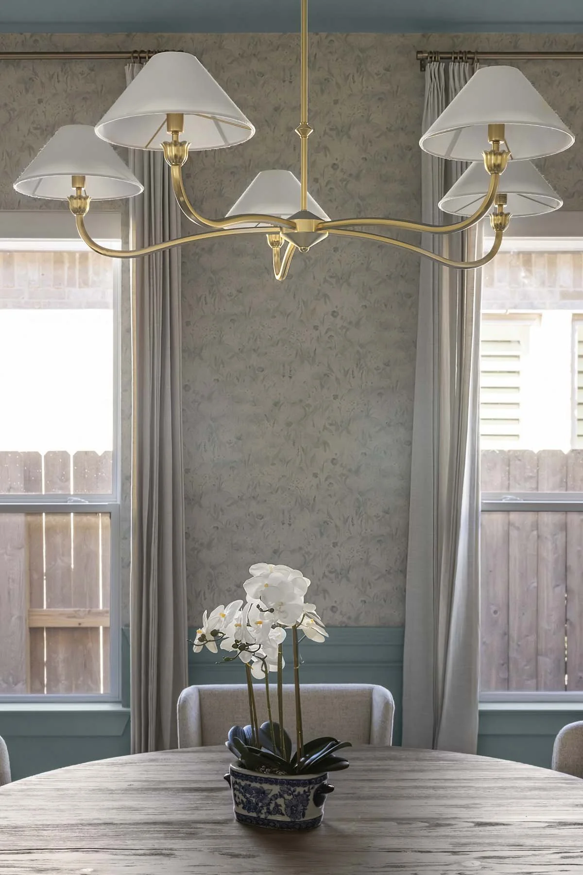 A dining room with a round wooden table, a white orchid in a decorative blue and white pot, a chandelier with five lampshades, large windows with curtains, and a wooden fence outside.