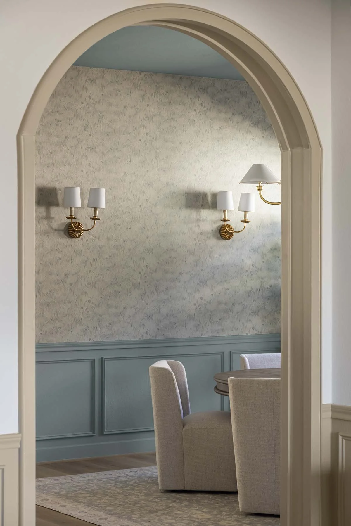 Interior view of a room with beige armchairs, a round wooden table, patterned wallpaper, and two wall-mounted brass light fixtures with white shades seen through an arched doorway.