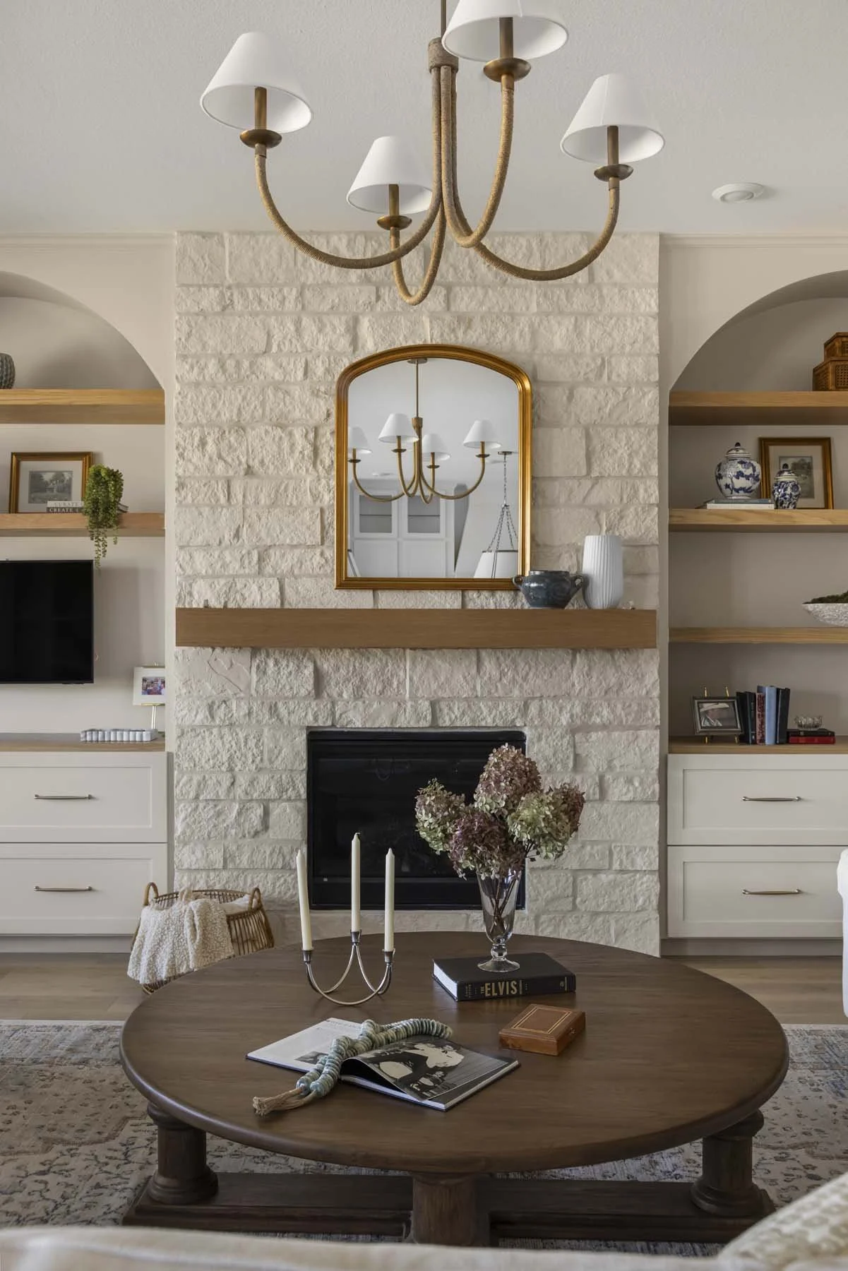 Living room with a white brick fireplace, wooden mantel, and built-in shelves. A round wooden coffee table with a flower vase, books, and decorative items sits in front. A chandelier hangs from the ceiling. A mirror reflects part of the room and the 