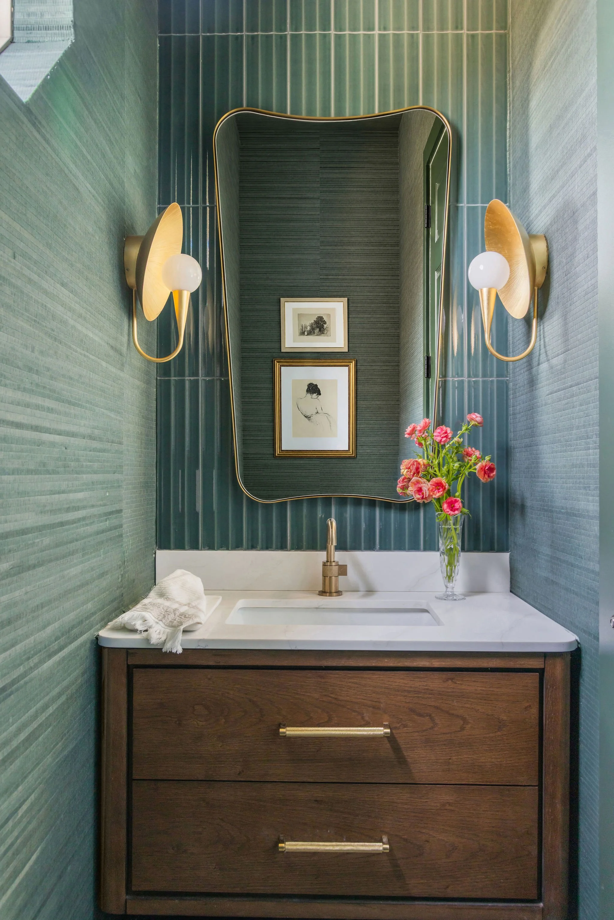 Modern bathroom vanity with a rectangular white sink, a gold faucet, a dark wood cabinet with gold handles, a large mirror with a gold border, two wall sconces with gold shades and white bulbs, a vase with pink flowers, and framed artwork on the wall