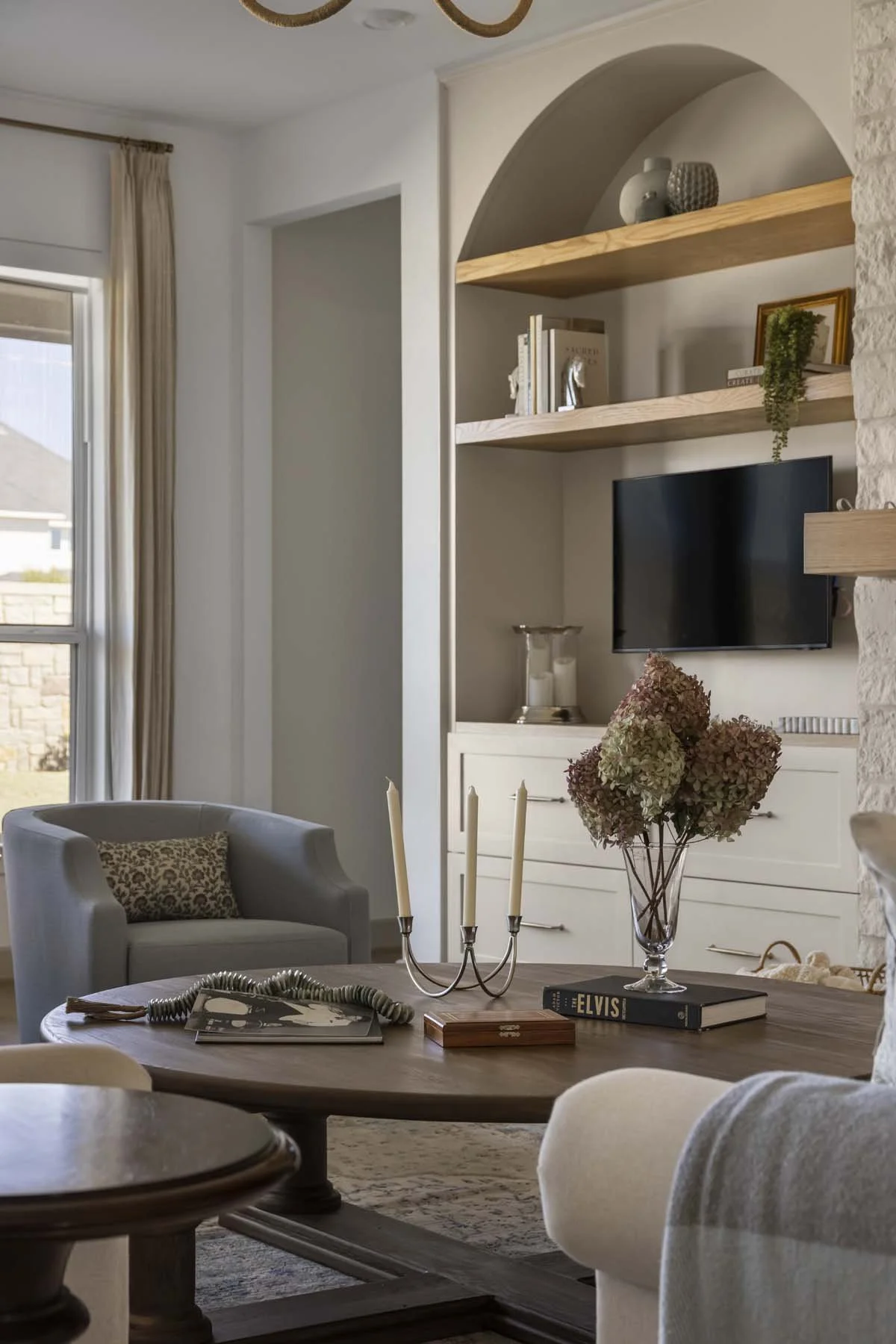Living room with a wooden dining table, a gray armchair with a patterned pillow, decorative books, a vase with dried flowers, a candelabra, a book titled "Elvis," and a mounted flat-screen TV.