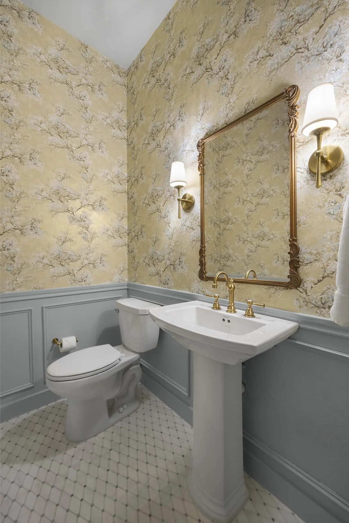 A vintage-style bathroom with a pedestal sink, a toilet, and a large mirror with gold accents. The walls have a floral-patterned wallpaper, and there are two wall sconces with white shades.