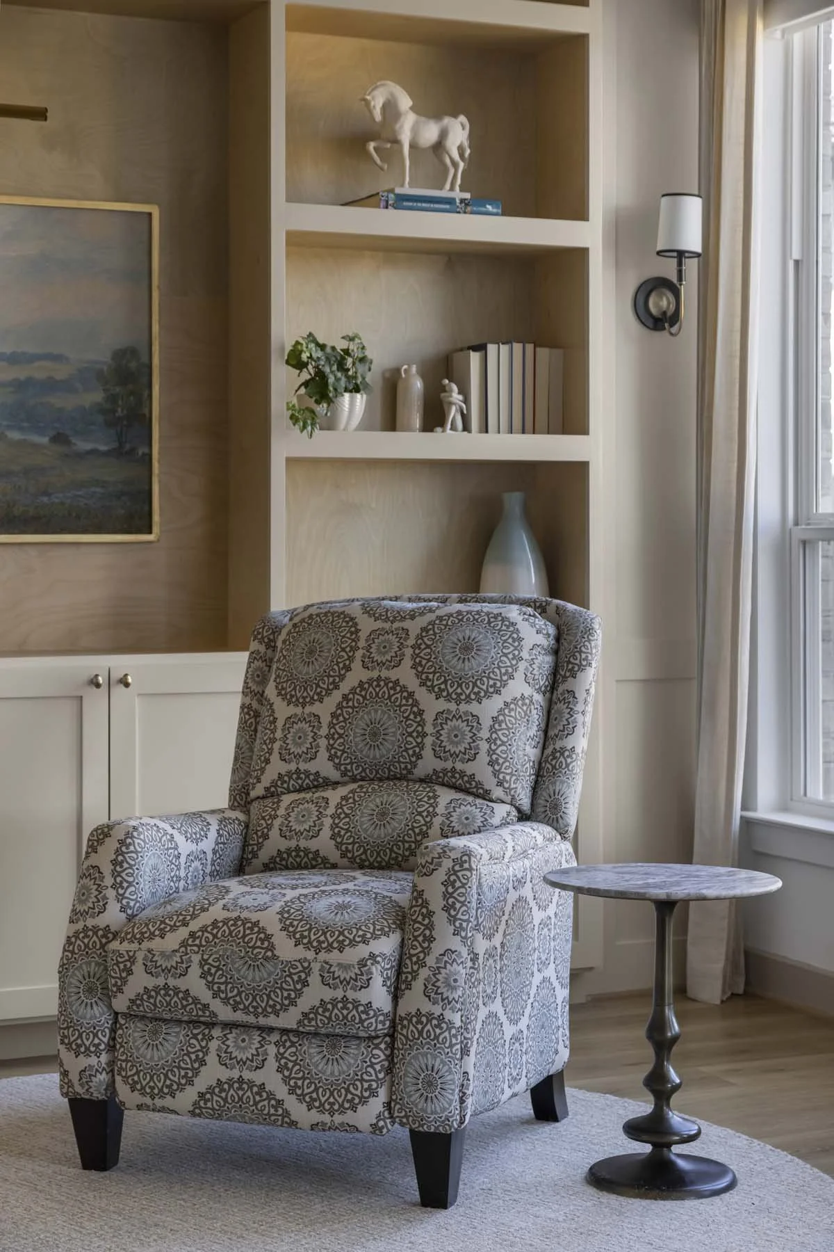A cozy living room corner with an armchair featuring a floral pattern, an accent table, built-in shelves with decorative items, and a large window with curtains.