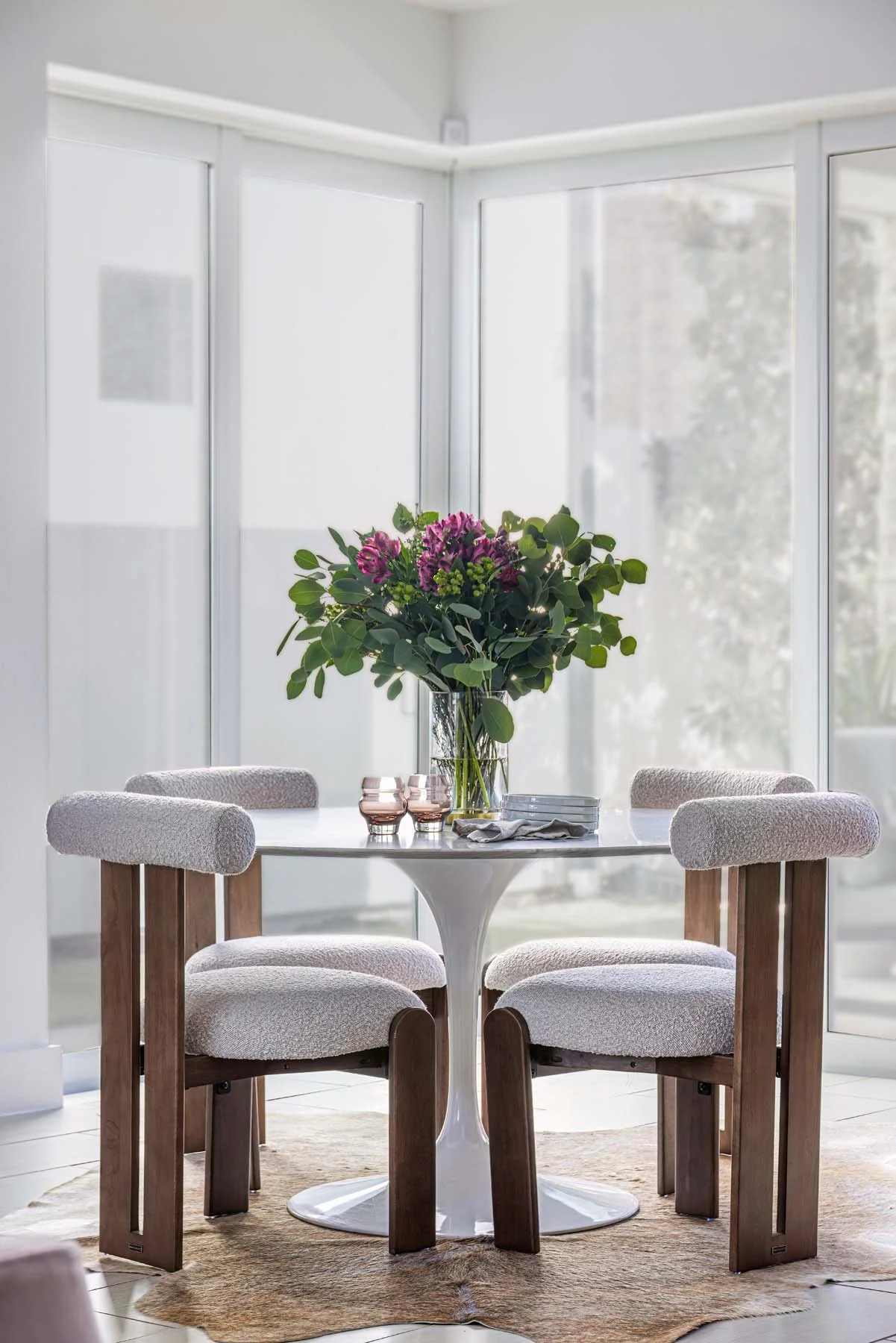 A round white dining table with a large bouquet of purple and green flowers, set with glasses, a bowl, a napkin, and plates, surrounded by four chairs with wooden supports and cushioned, textured beige seats, in front of large windows letting in natu