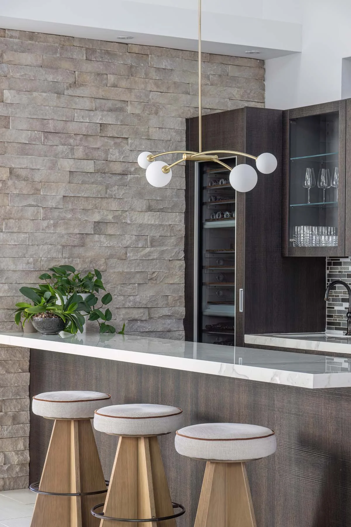 Modern kitchen with stone accent wall, white marble countertop, three wooden barstools with upholstered seats, a brass and white globe chandelier, a plant, and a wine cooler.