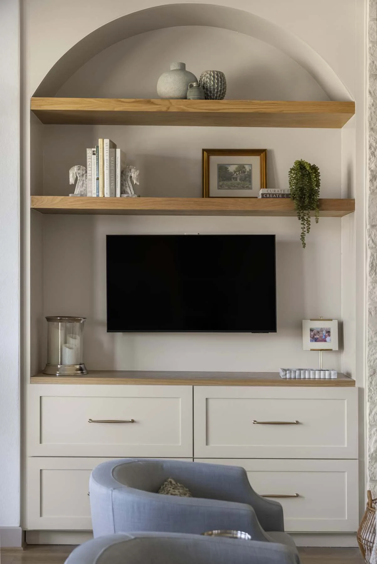 Living room with a built-in media console, mounted flat-screen TV, wooden shelves with decorative objects, books, framed art, and a small table lamp, with a blue armchair in the foreground.