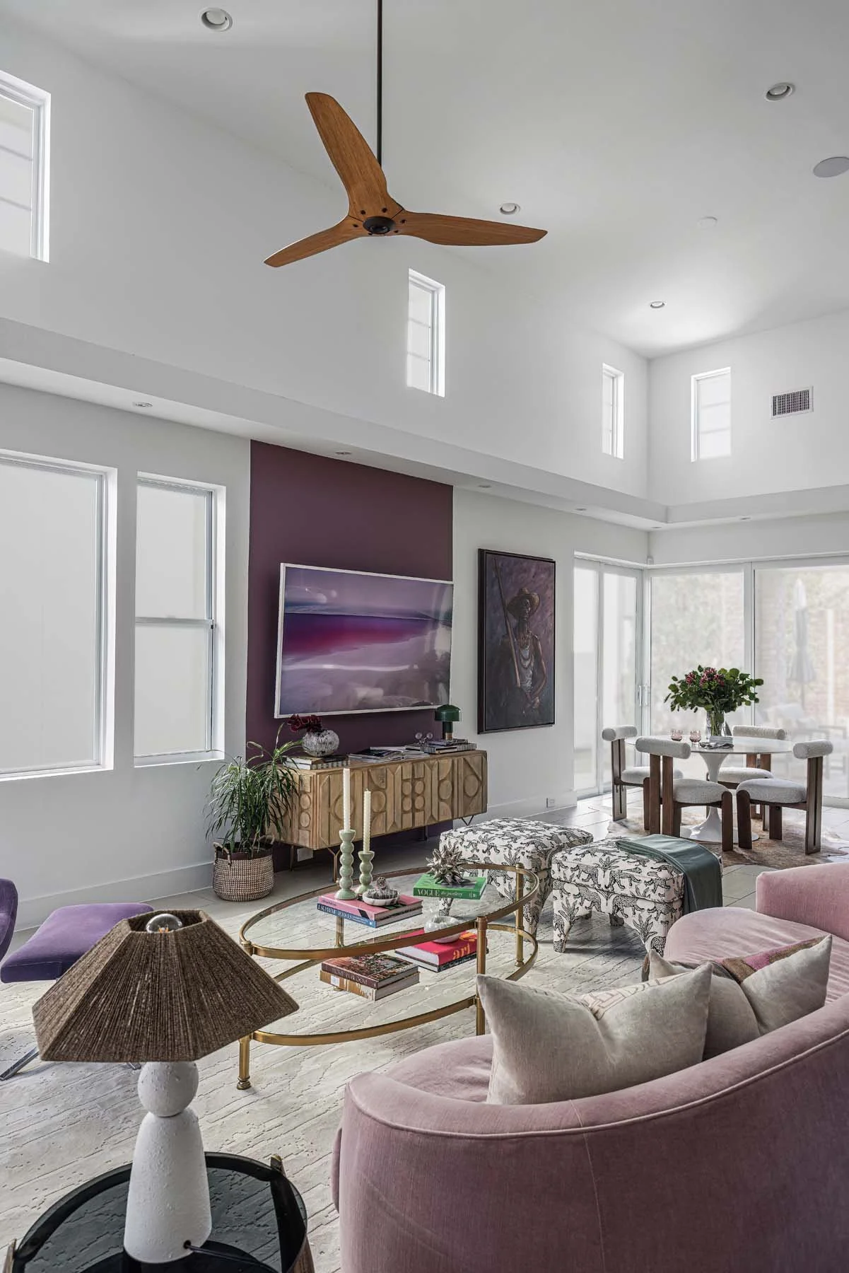 Bright and modern living room with high white ceiling and multiple tall windows letting in natural light, ceiling fan with wooden blades, a purple accent wall with a flat-screen TV, a wooden sideboard, a round dining table with chairs, pink upholster